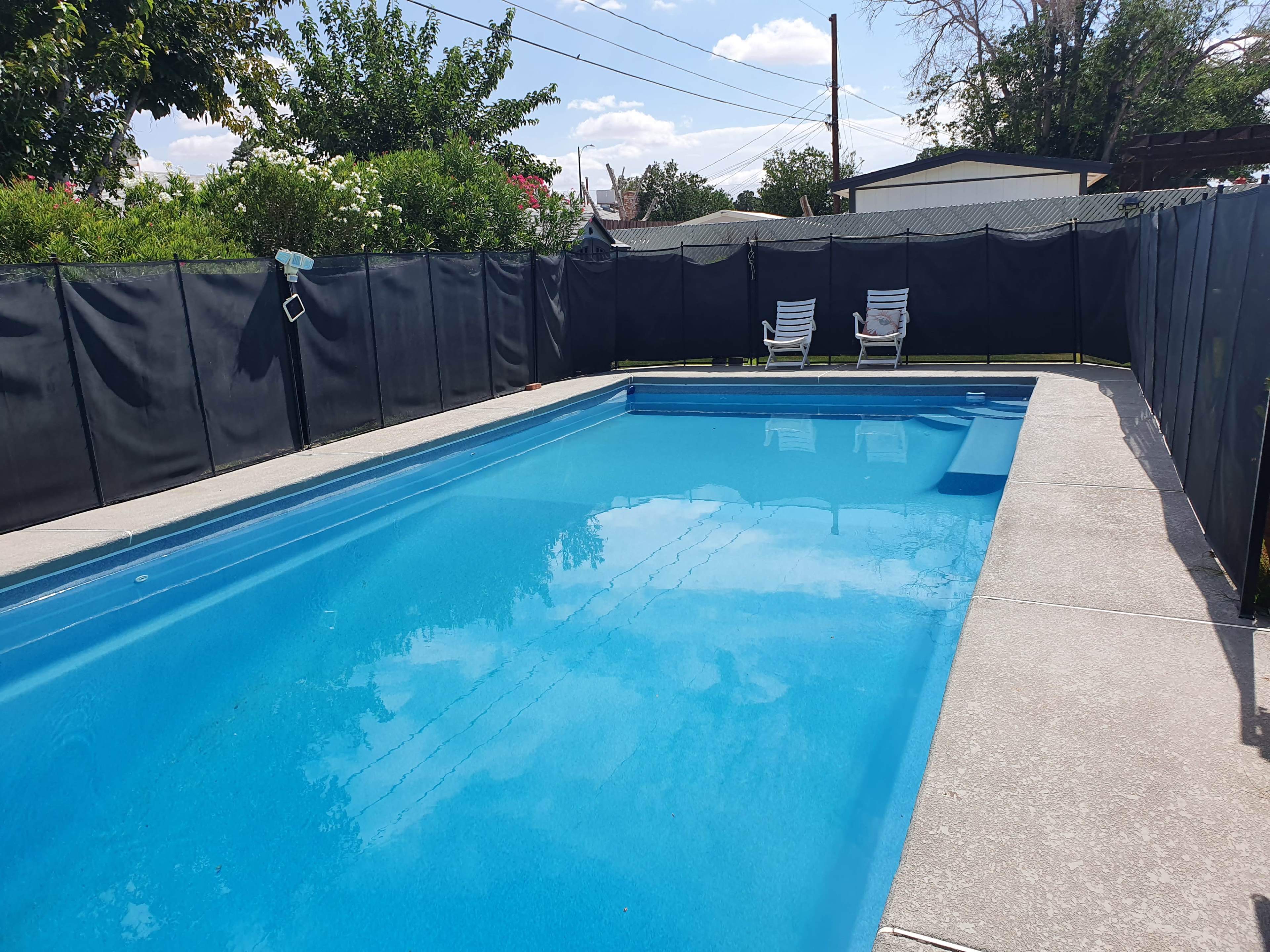 A clear swimming pool is surrounded by a black fence, with two lounge chairs positioned on the deck.