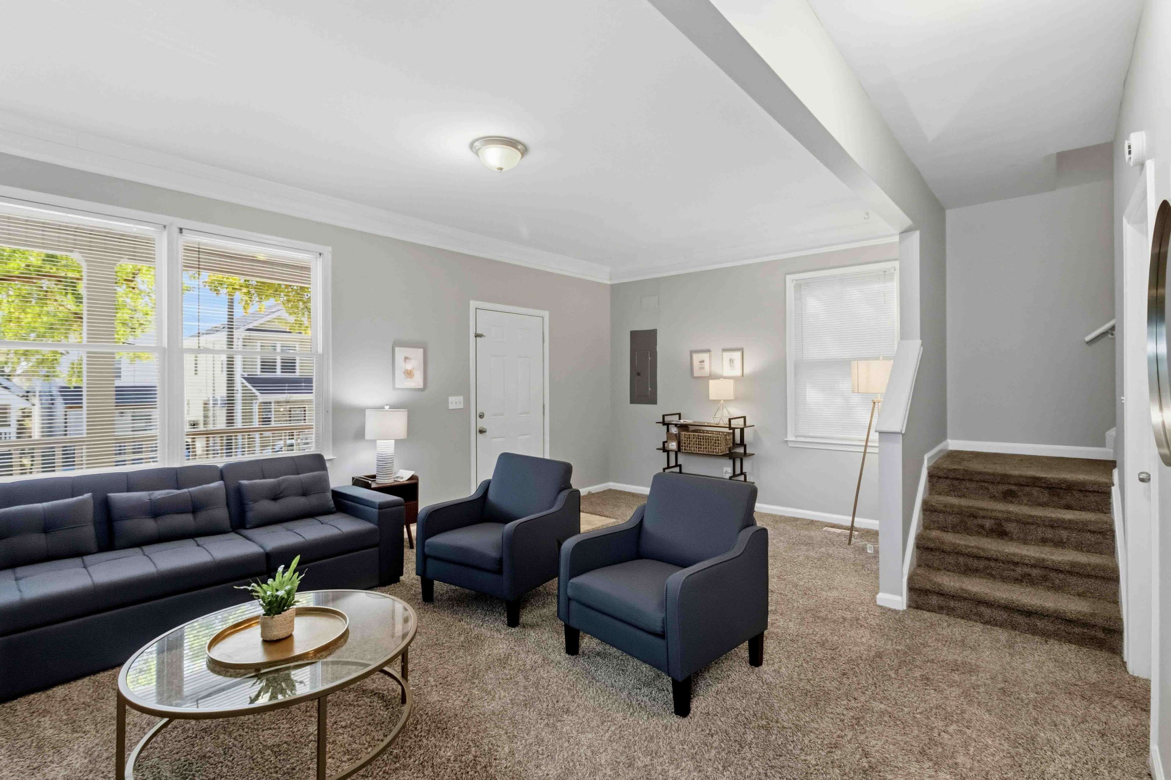 The image depicts a living room featuring a dark sofa, two armchairs, a round coffee table, and a staircase in a well-lit space with large windows.
