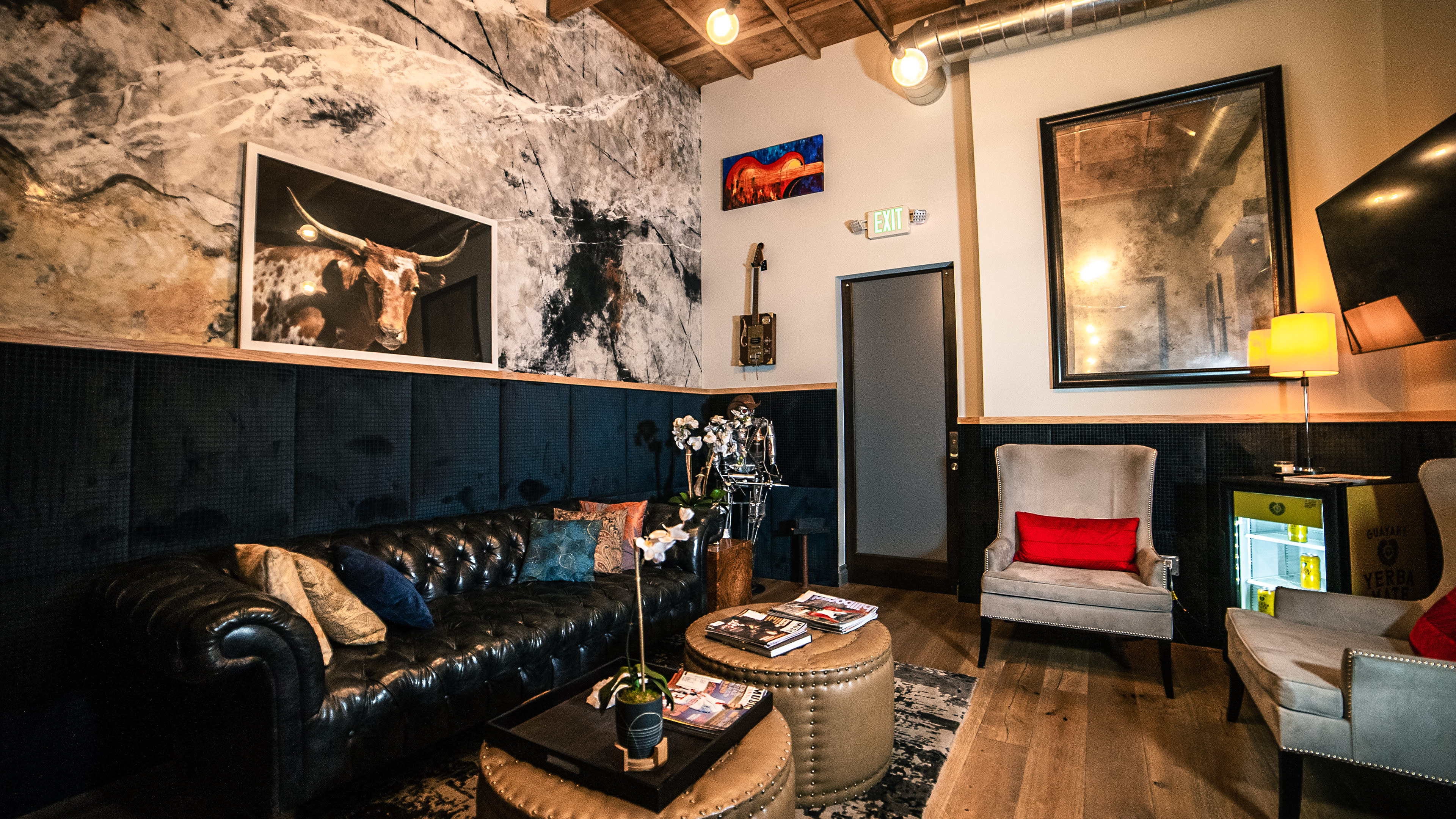 The image shows a stylish lounge with a dark leather sofa, a circular coffee table, and various artworks adorning the walls.