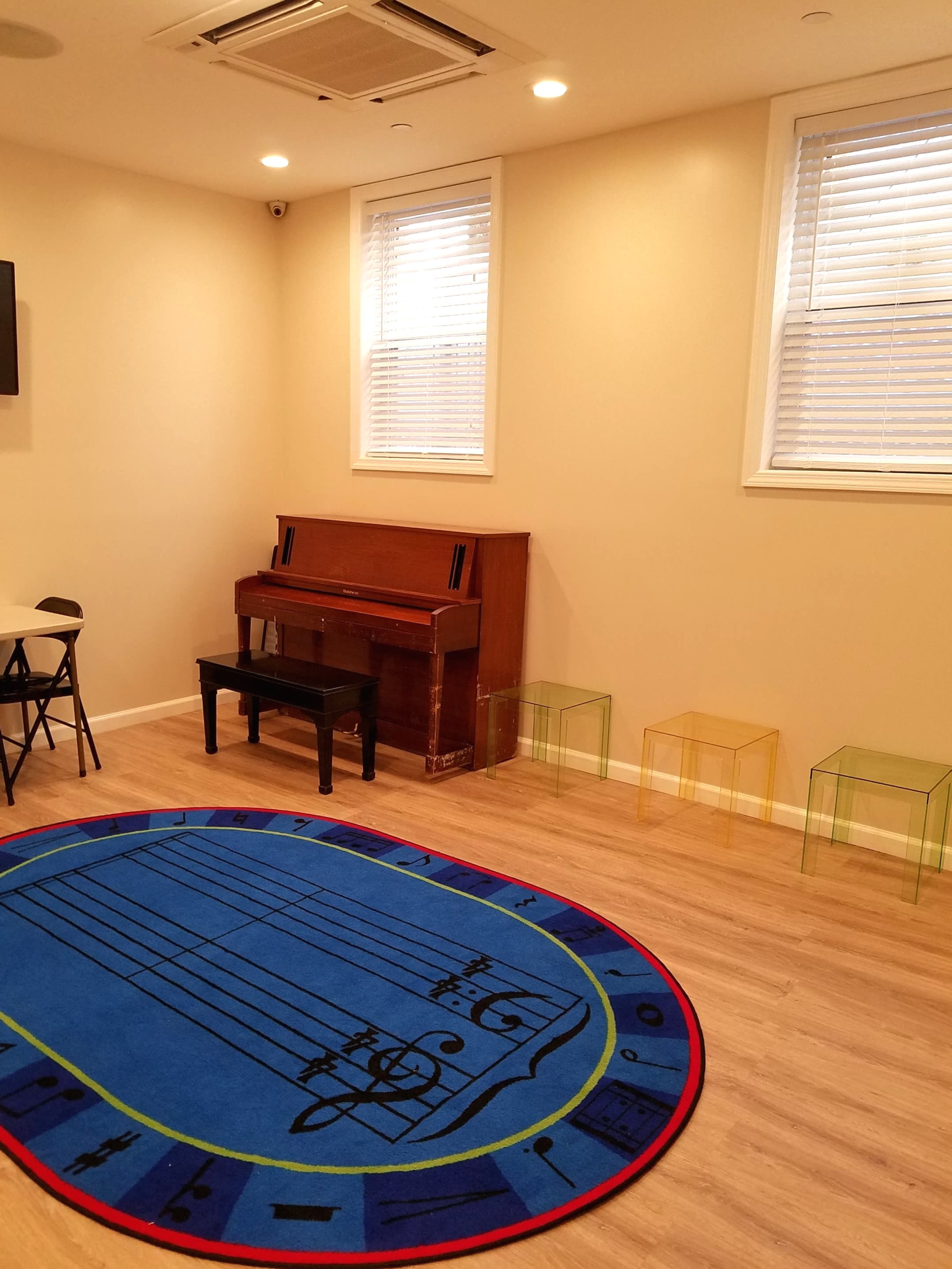 A room with light-colored walls contains a piano, colorful transparent chairs, and a blue area rug featuring musical notes.