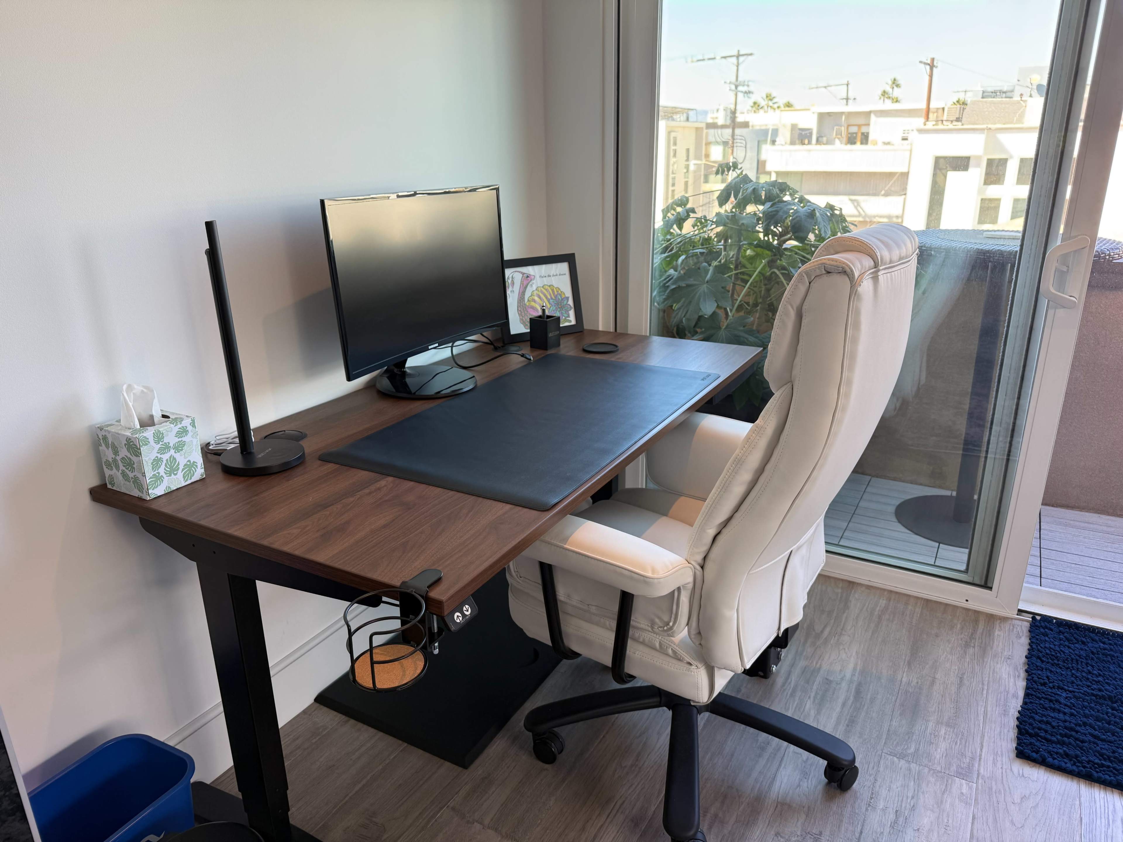 The image shows a modern workspace featuring a wooden desk with a computer monitor, a black desk mat, a task lamp, and an office chair, situated next to a large window with a view of a balcony and greenery outside.