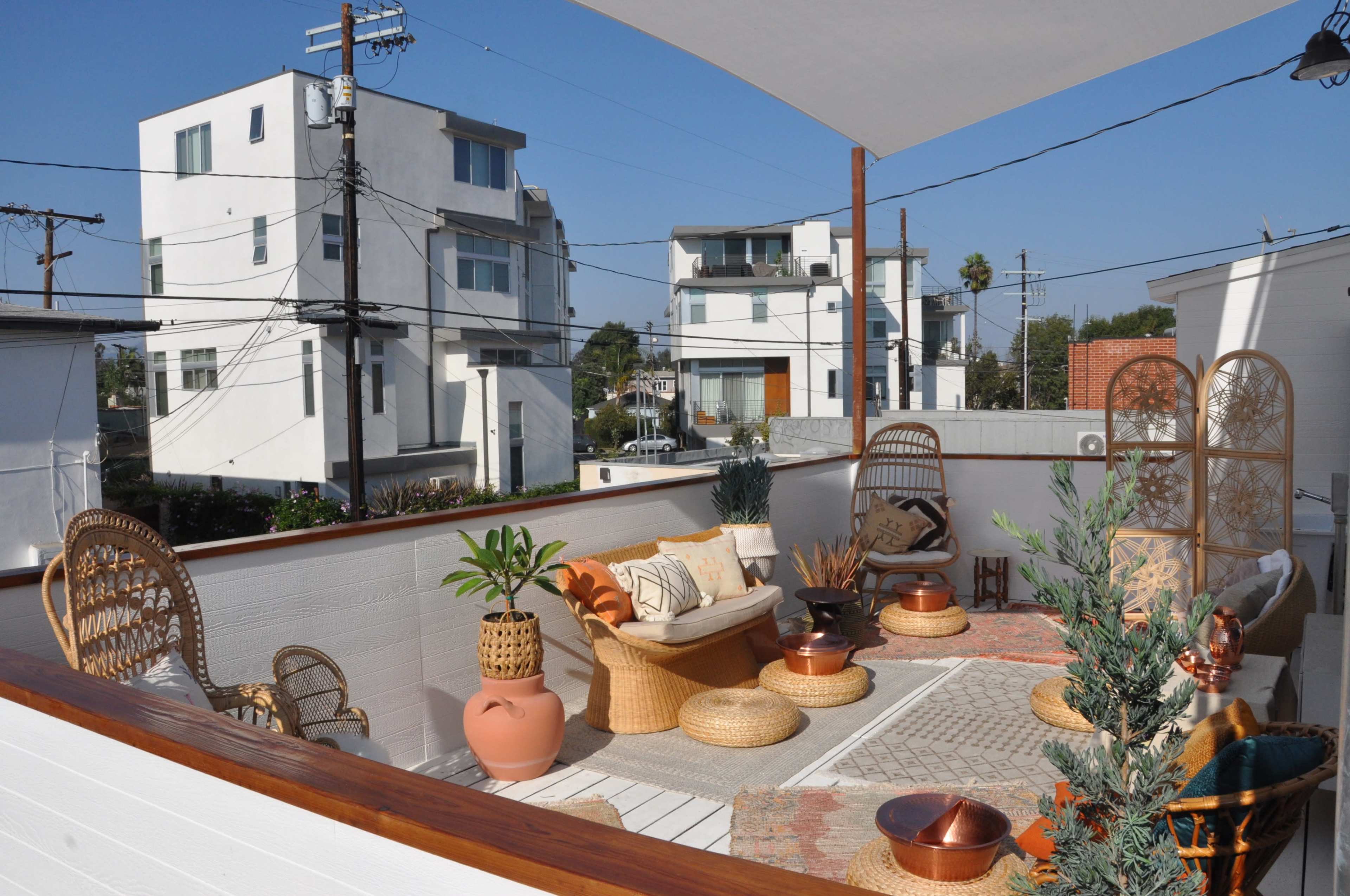 The image shows a modern rooftop patio decorated with wicker furniture, potted plants, and a neutral rug, surrounded by contemporary buildings and utility lines.