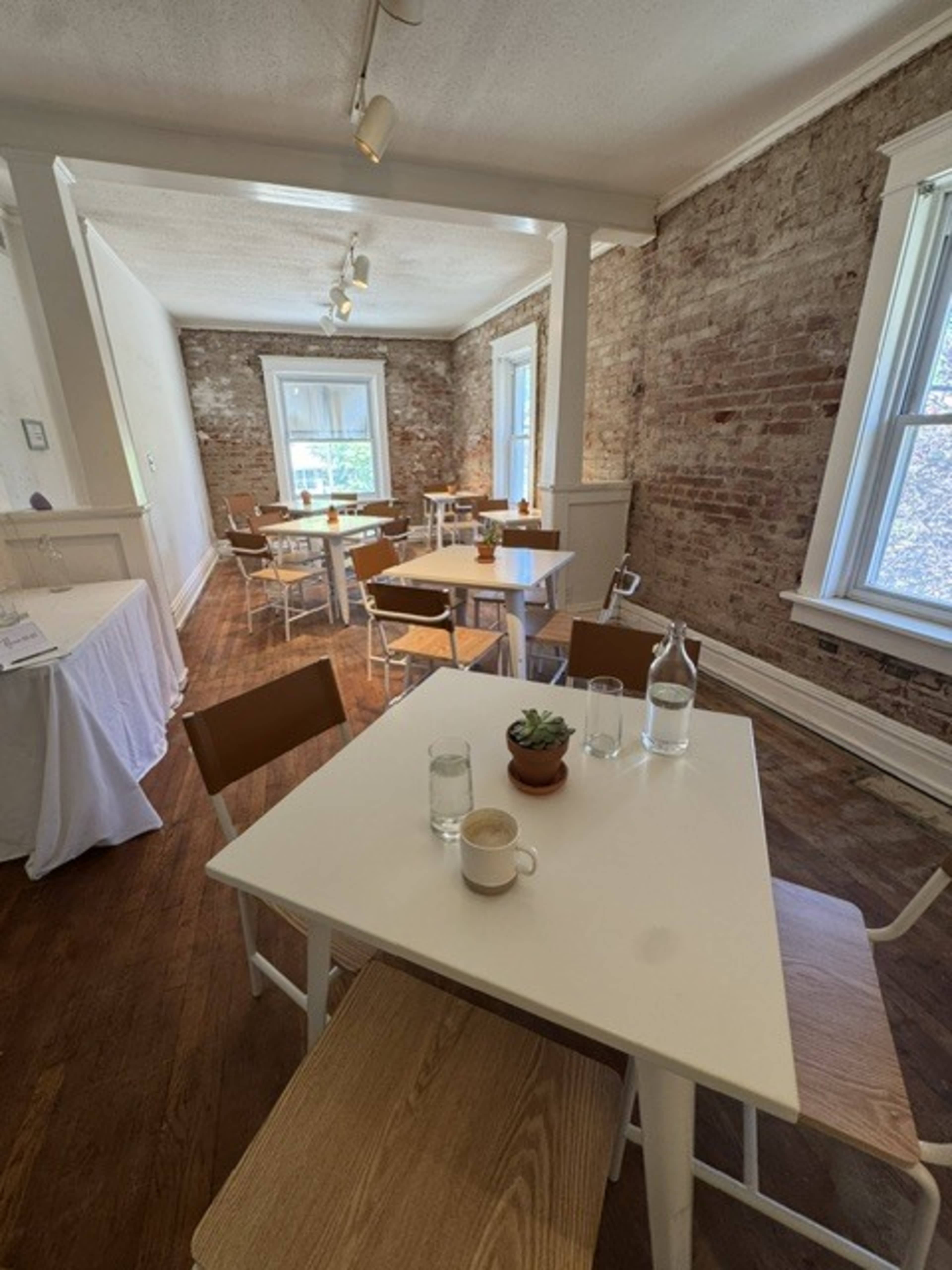 The image shows a rustic, well-lit café with wooden floors, exposed brick walls, and several tables and chairs arranged throughout the space.