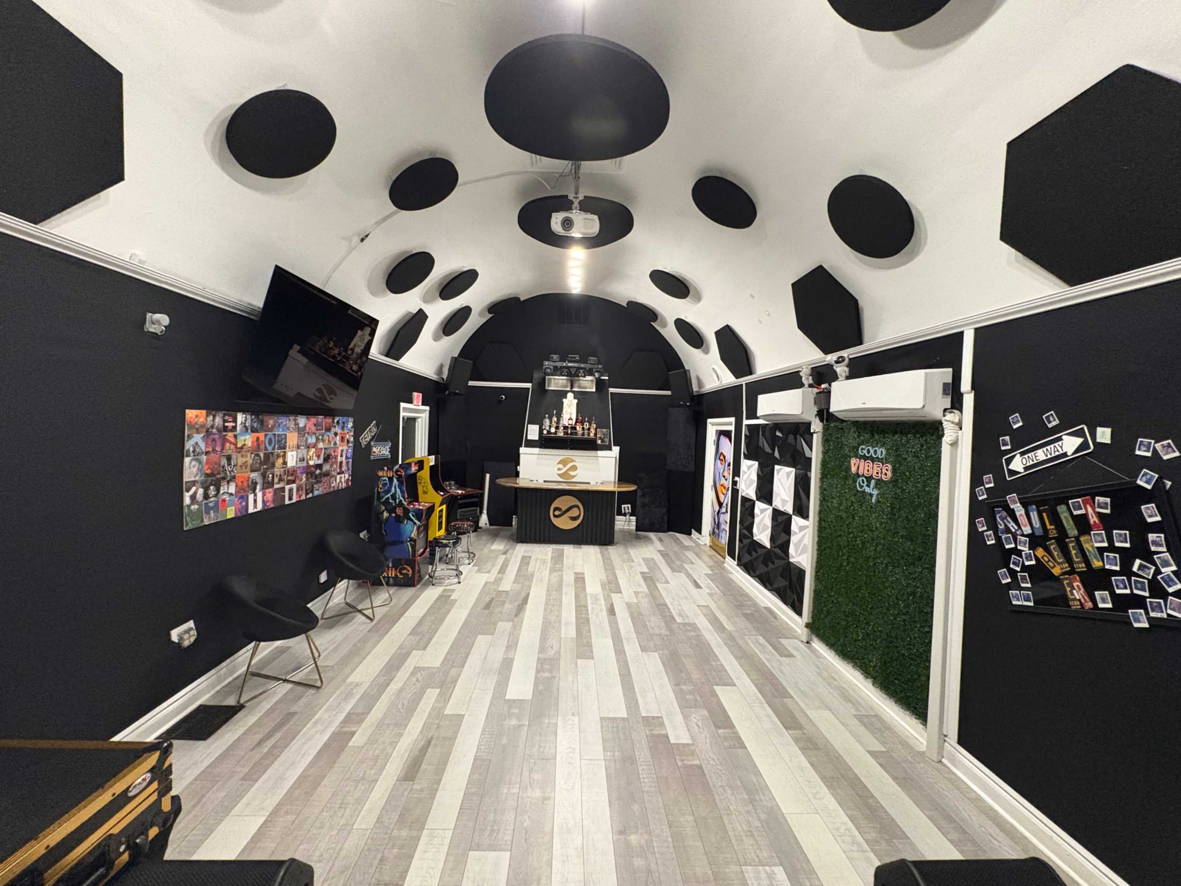 The image shows a modern interior space with black walls, a bar area at the far end, hexagonal ceiling panels, and decorative artwork on the walls.