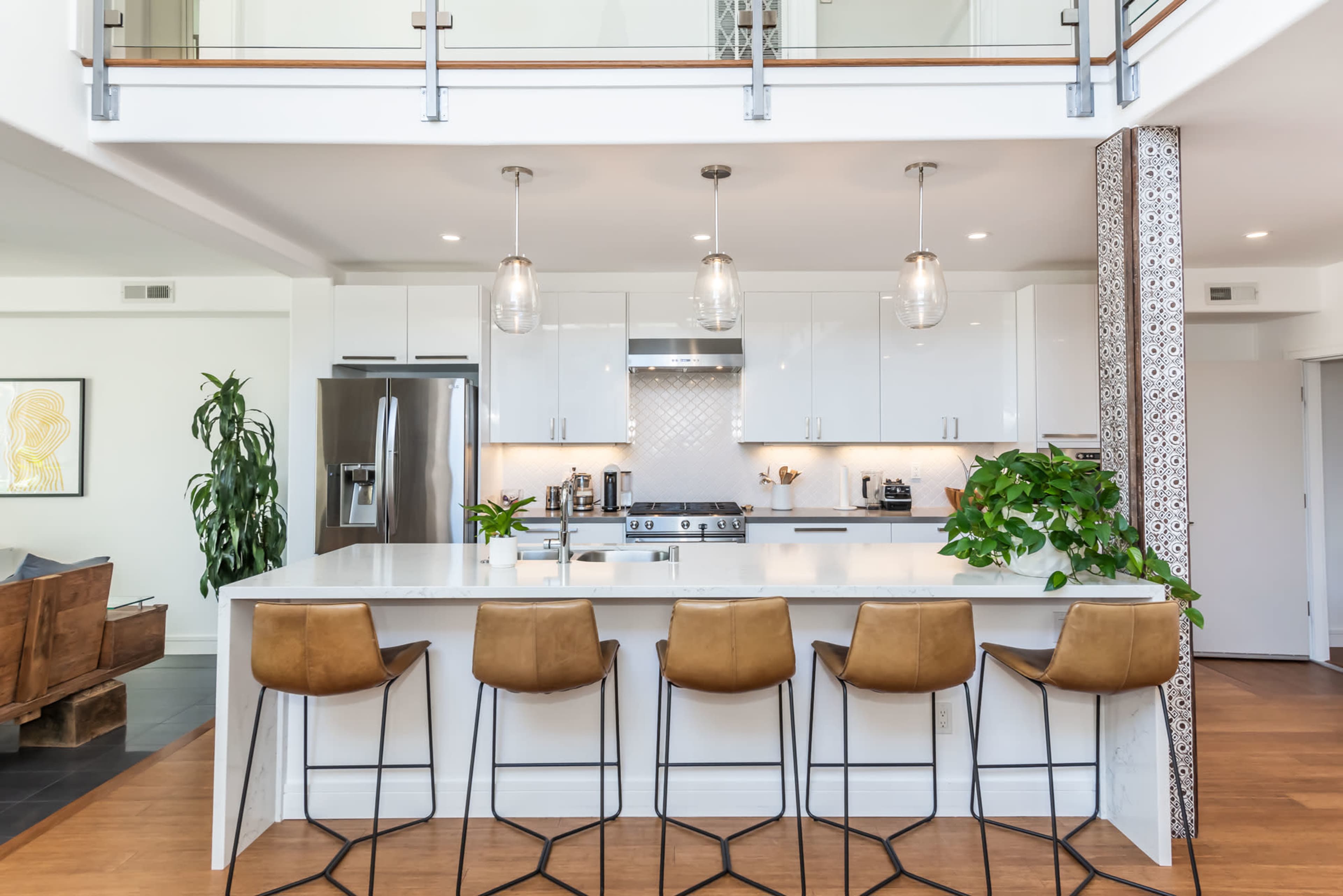 A modern kitchen with a white island counter, three pendant lights, and bar stools, situated in an open floor plan with plants and stainless steel appliances.