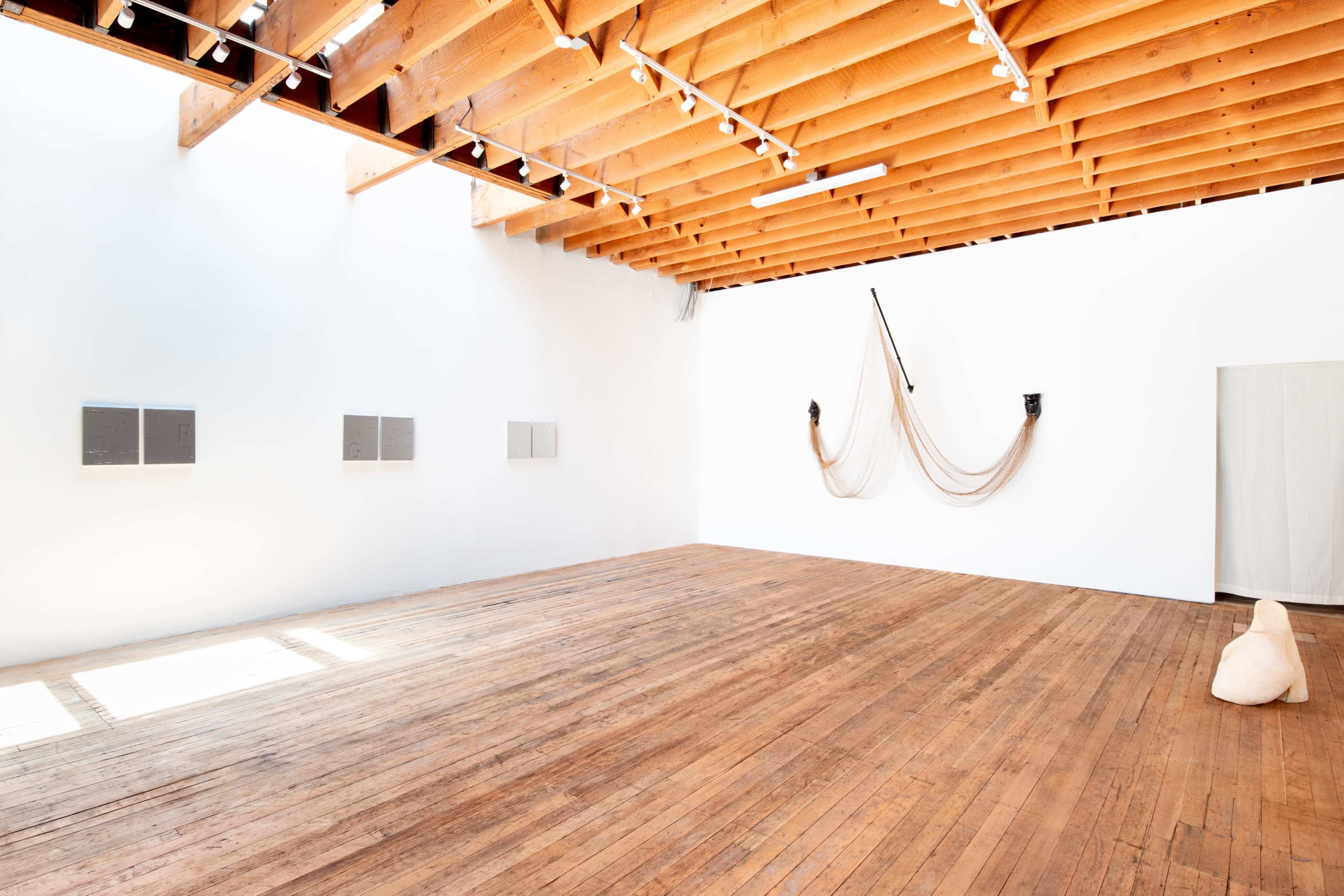 A minimalist art gallery with wooden flooring, white walls, and two distinct installations: a draped object with two black masks and several panels displayed on the left side.