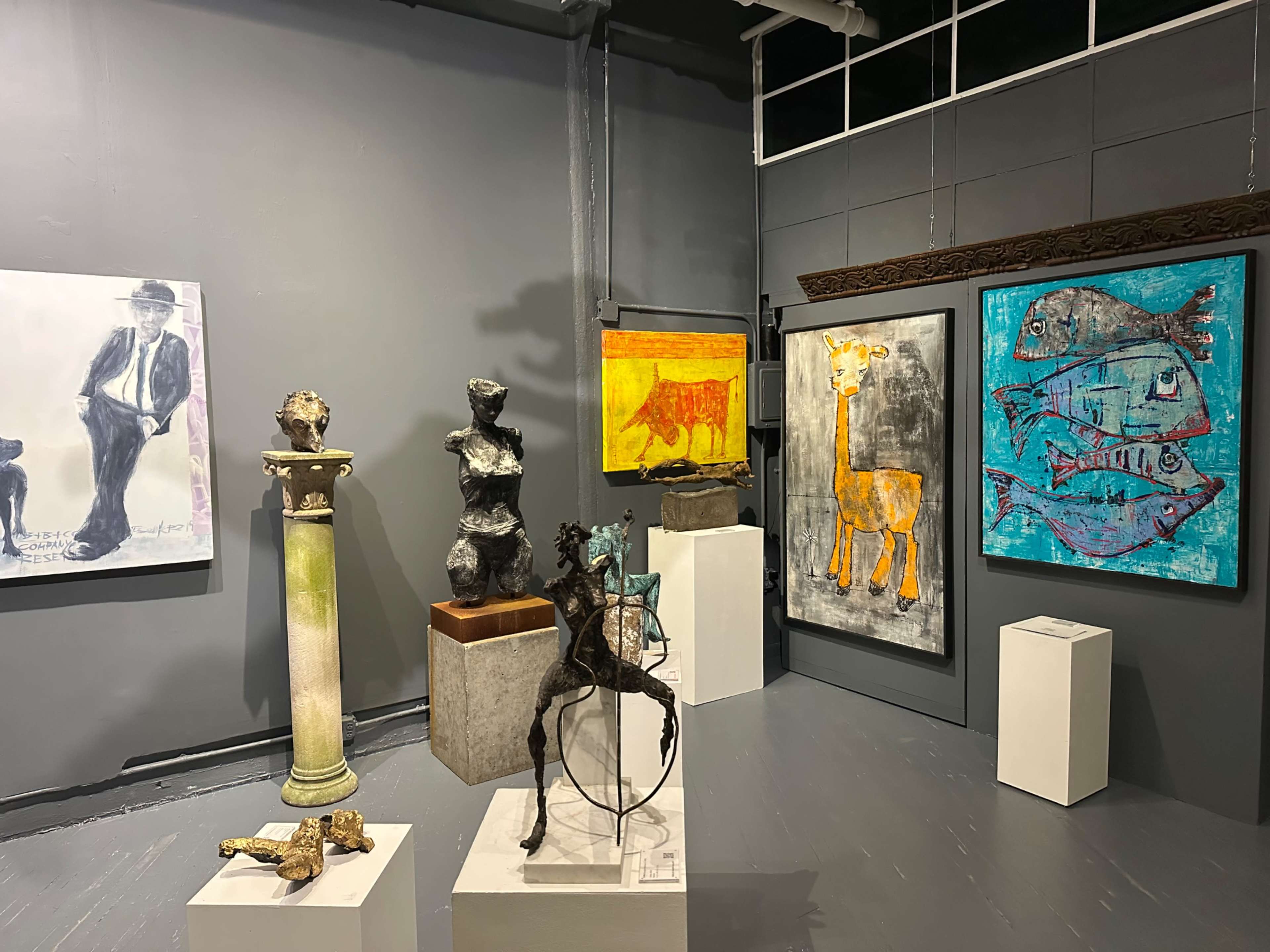The image shows an art gallery featuring various sculptures and paintings displayed against gray walls.