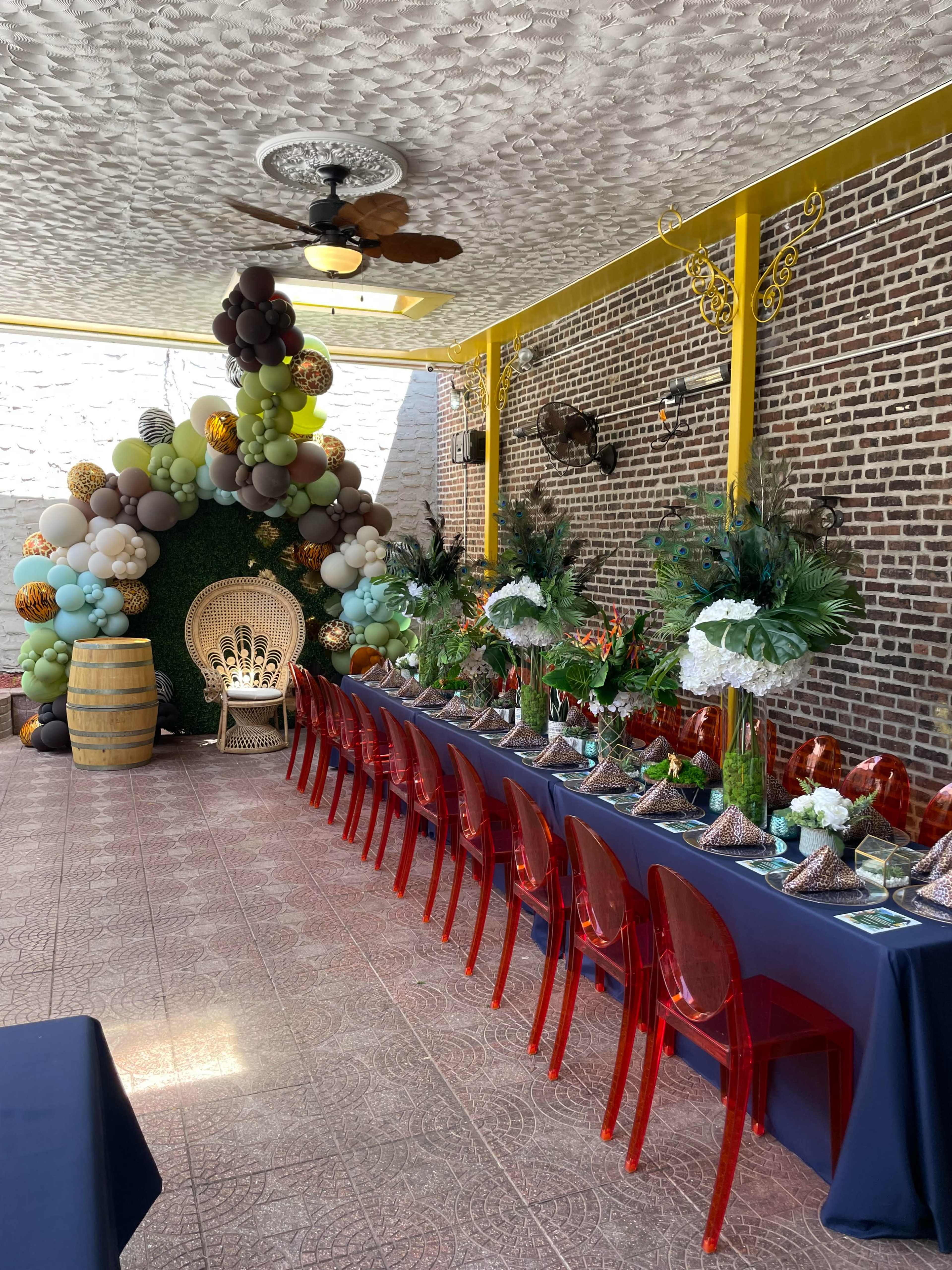The image shows a decorated indoor space featuring a long table set with floral arrangements and party hats, alongside a balloon arch and a large decorative chair in a corner.