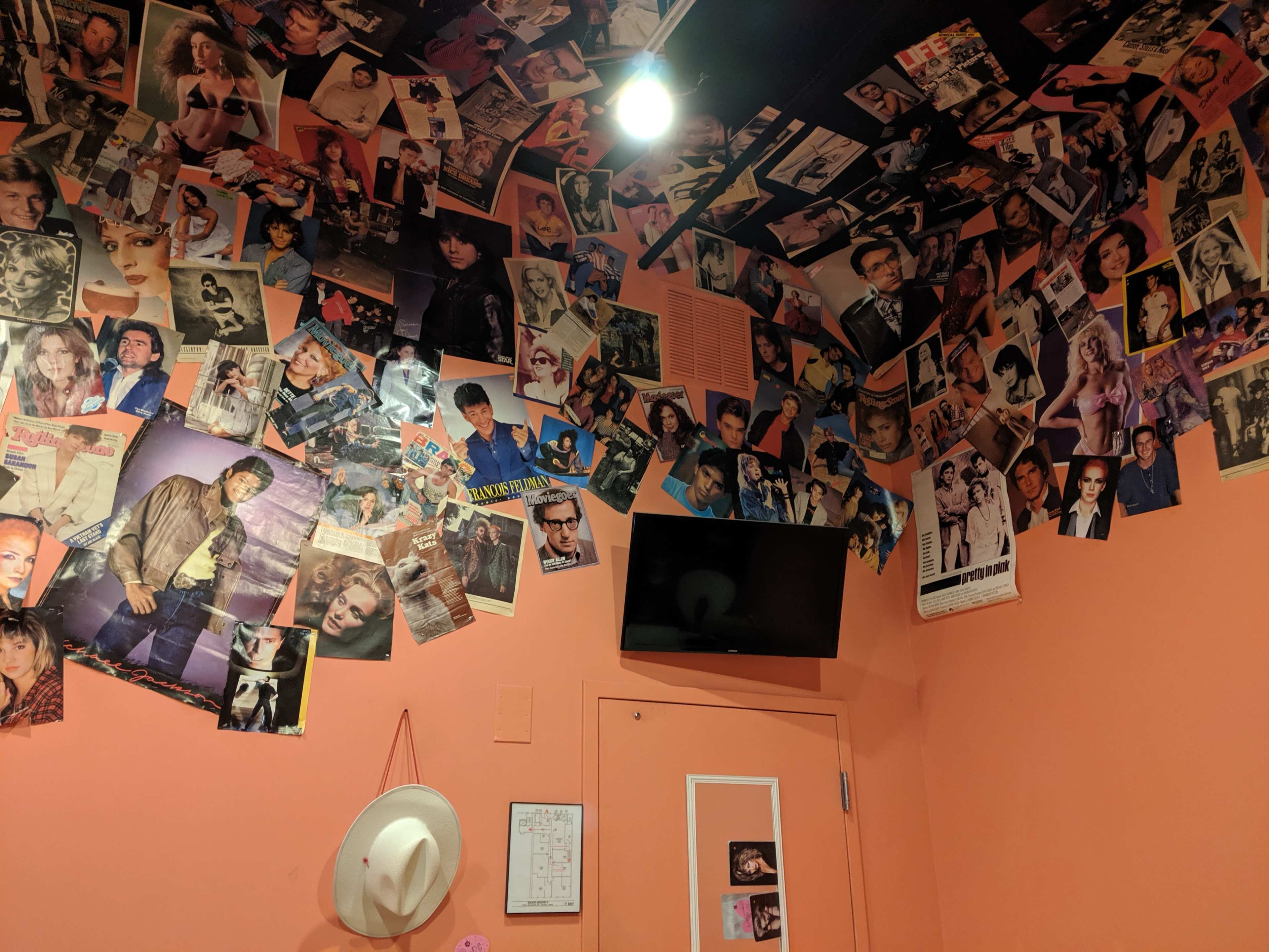 A room with walls covered in a collage of various celebrity posters and photographs, with a television mounted on one wall and a white hat hanging nearby.