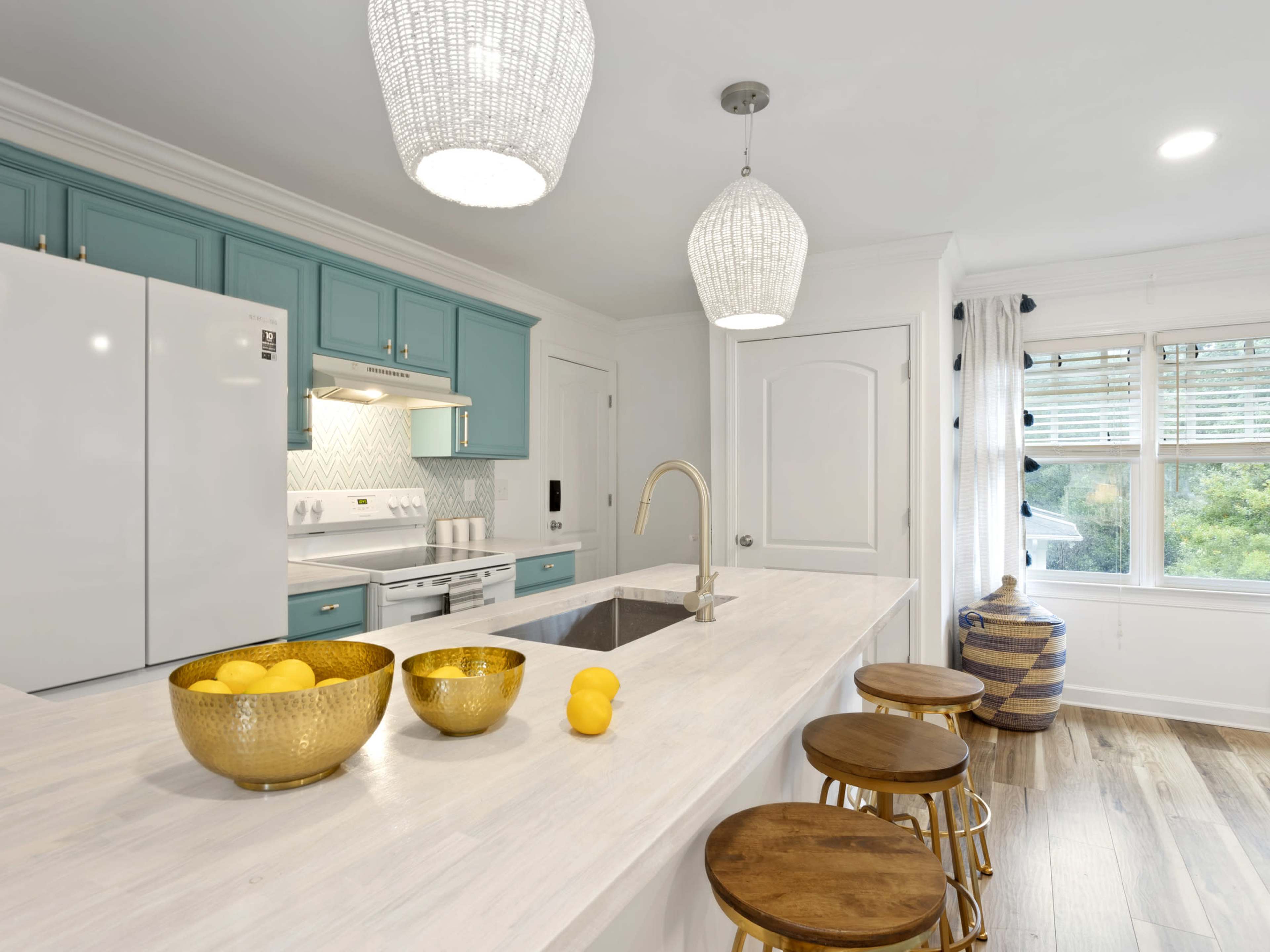 A modern kitchen features blue cabinetry, a white countertop with bowls of lemons, and pendant lighting above a central island with wooden stools.