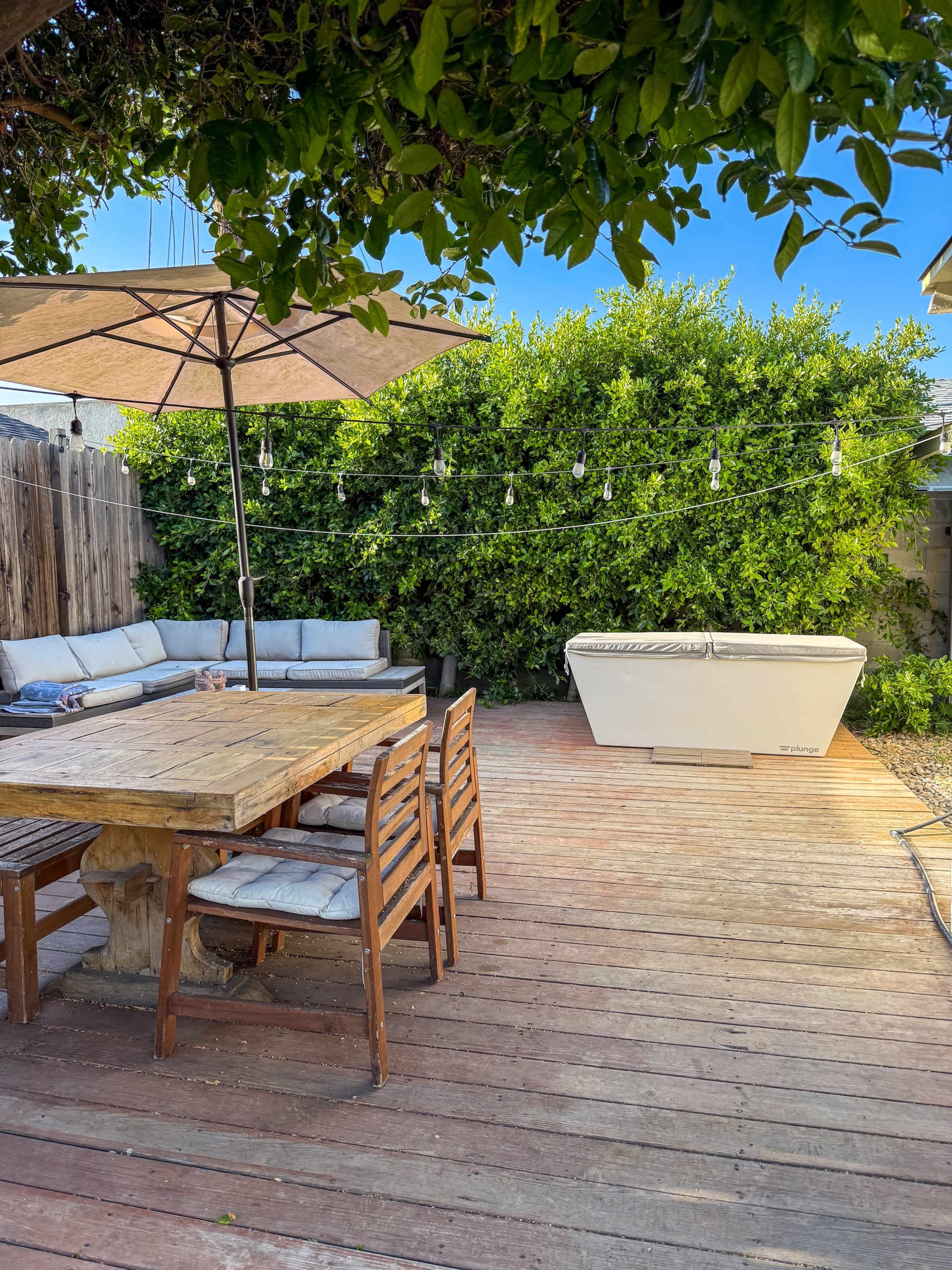 The image shows a wooden deck with a dining table and chairs, an umbrella, a lounge area, and a hot tub, surrounded by greenery.