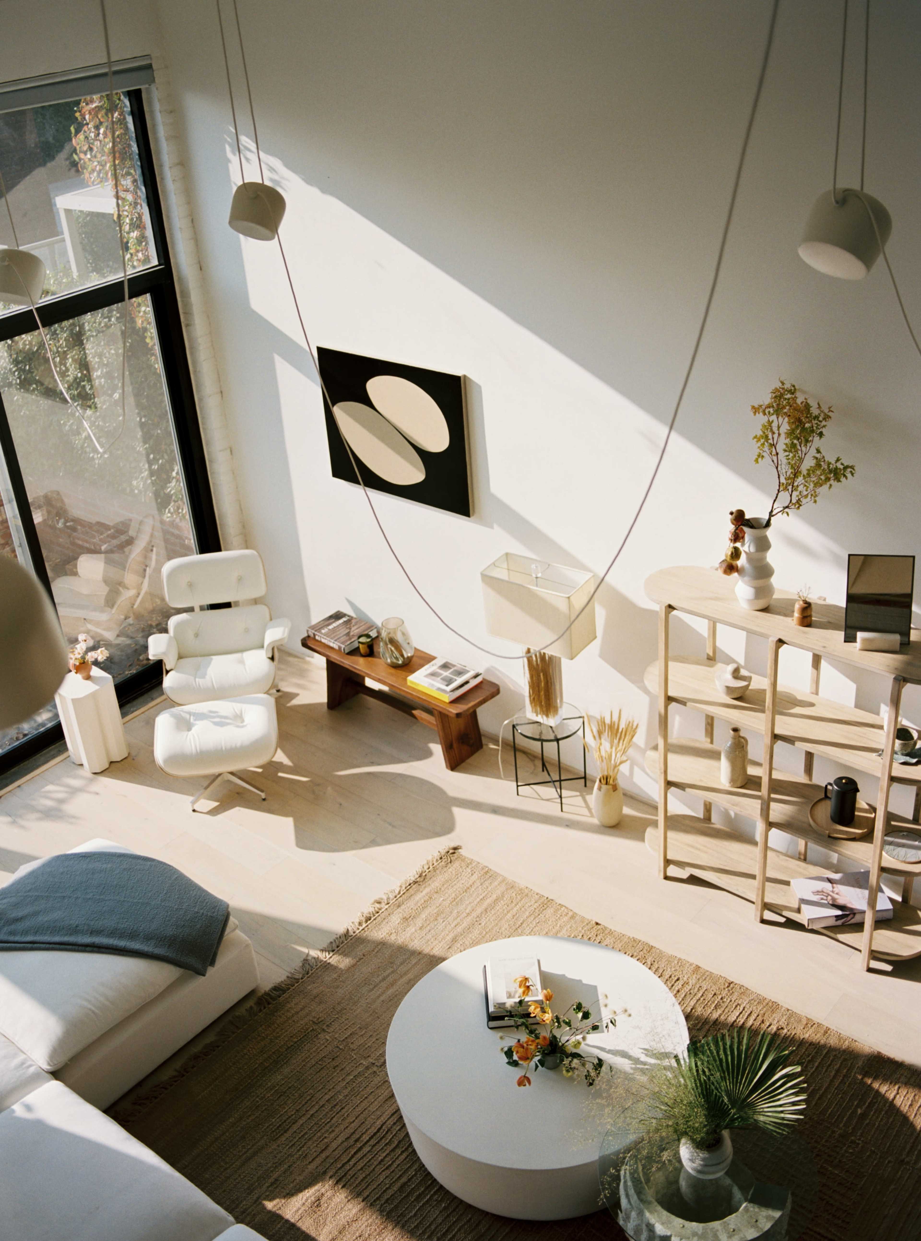 A minimalist living room features a circular coffee table, a woven rug, a modern chair, and shelves displaying decorative items, all illuminated by natural light from large windows.