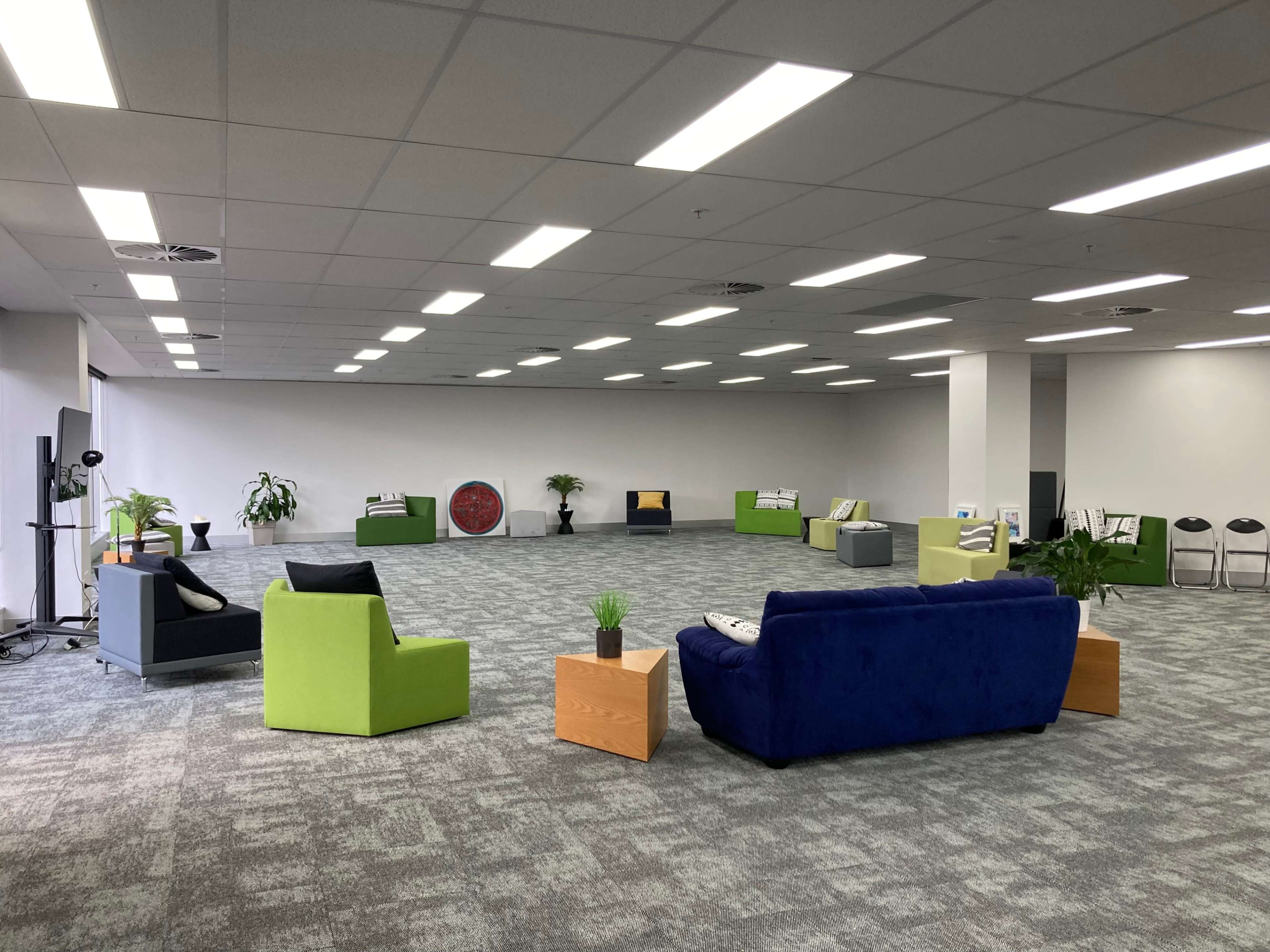 The image shows a spacious, brightly lit room with various seating arrangements consisting of colorful chairs and sofas, along with decorative plants placed around the area.