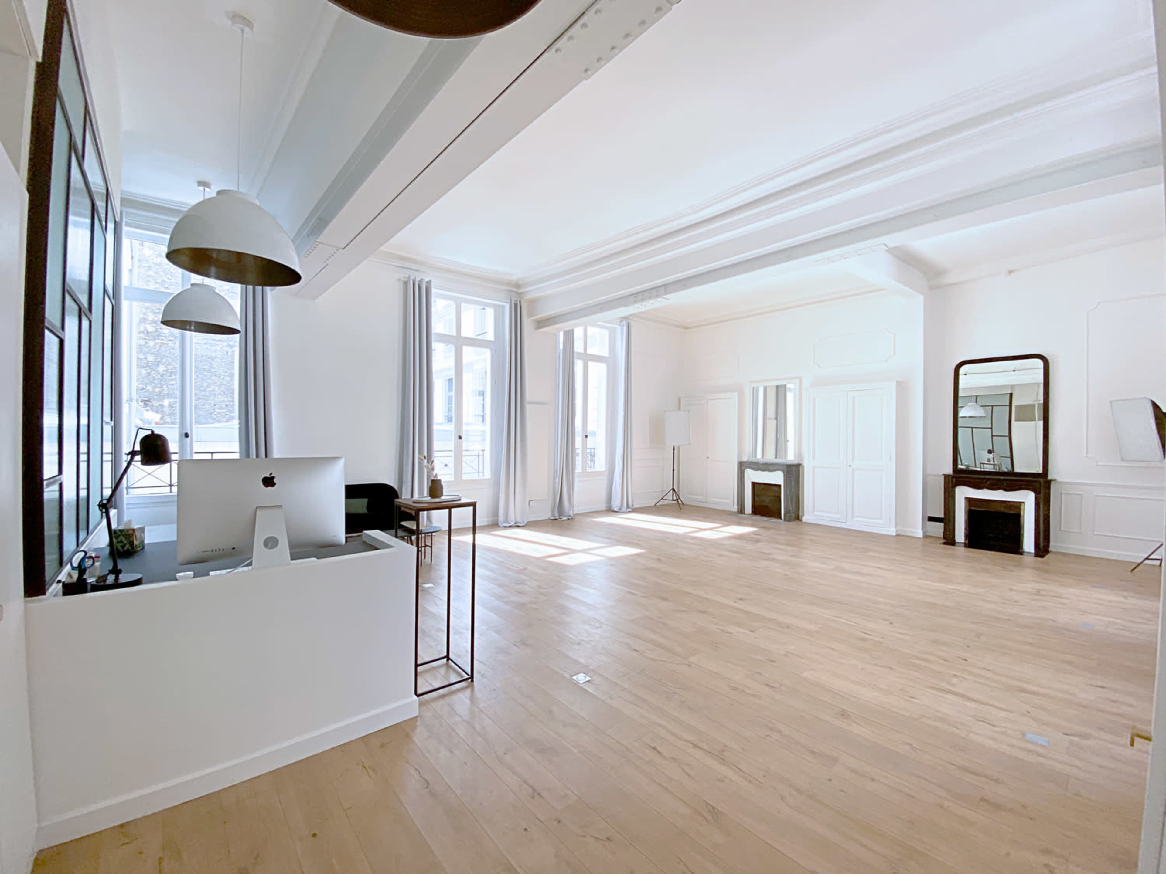 A bright, spacious room with light wooden flooring, large windows, and minimalist furniture, including a desk with a computer and a mirror.