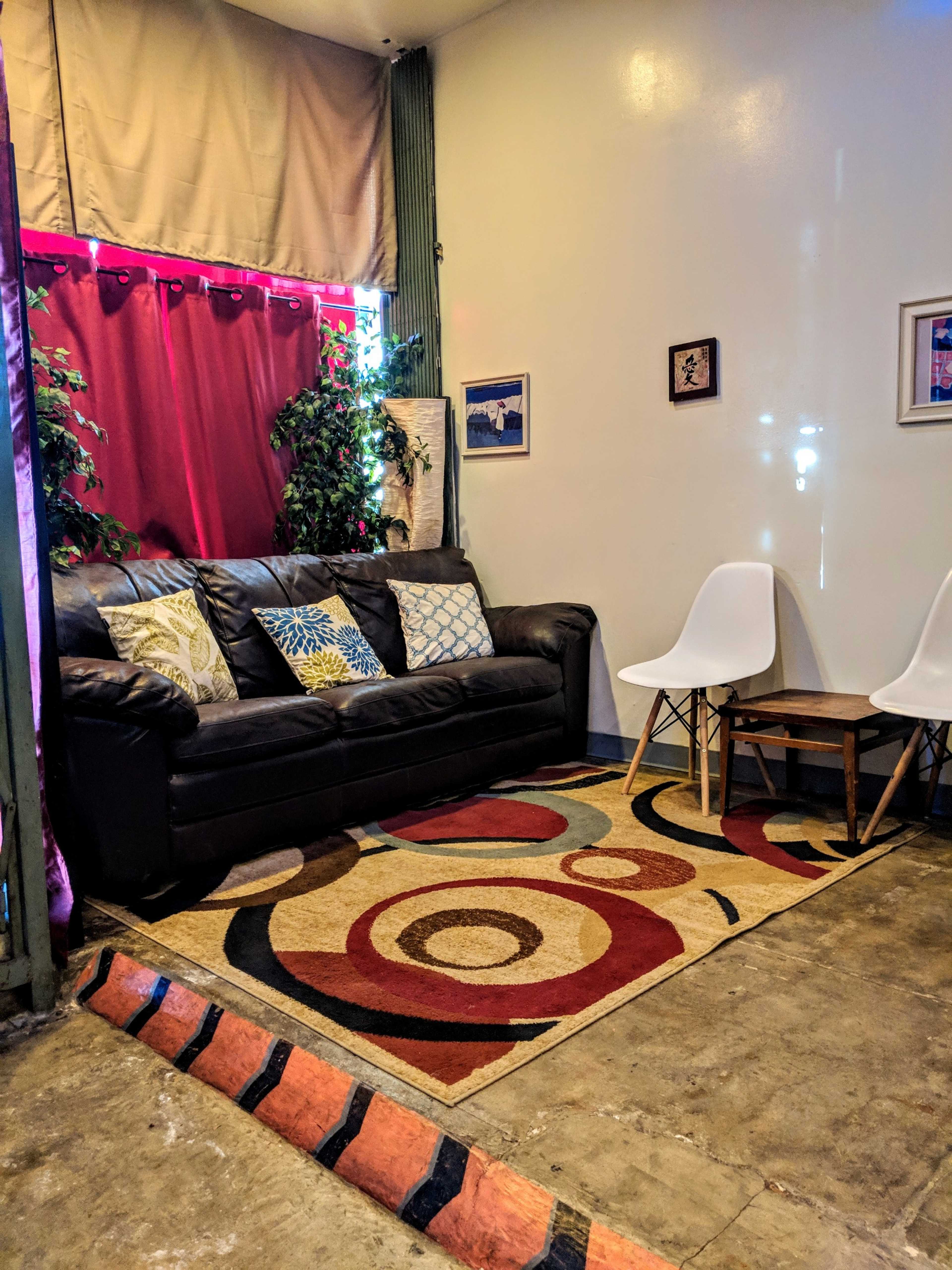 A small seating area with a black couch, white chairs, and a colorful rug on a concrete floor, surrounded by plants and decorations.