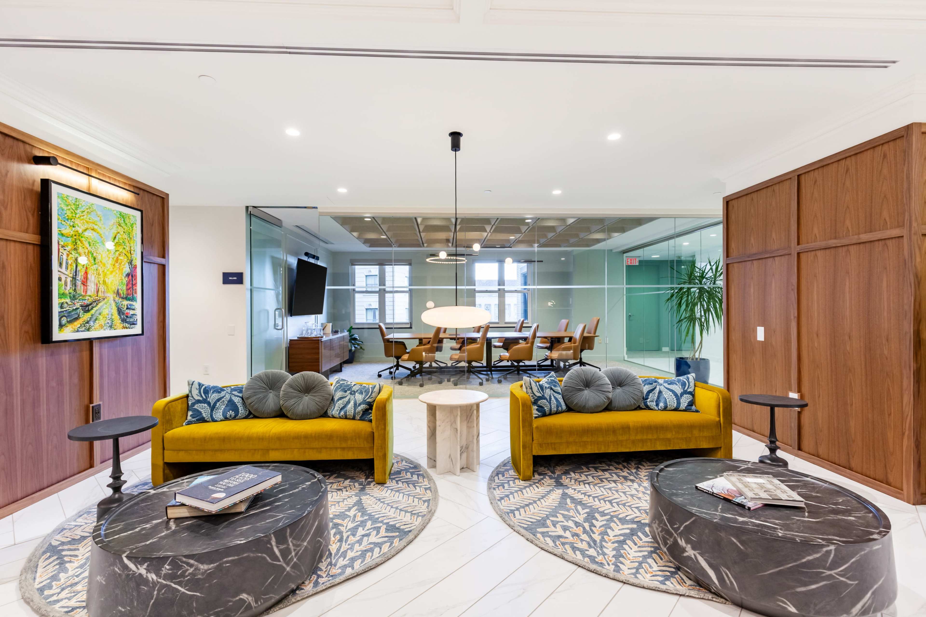 The image shows a modern office lobby featuring two yellow sofas with decorative pillows, two dark circular coffee tables, and a glass meeting room with a large conference table and chairs in the background.