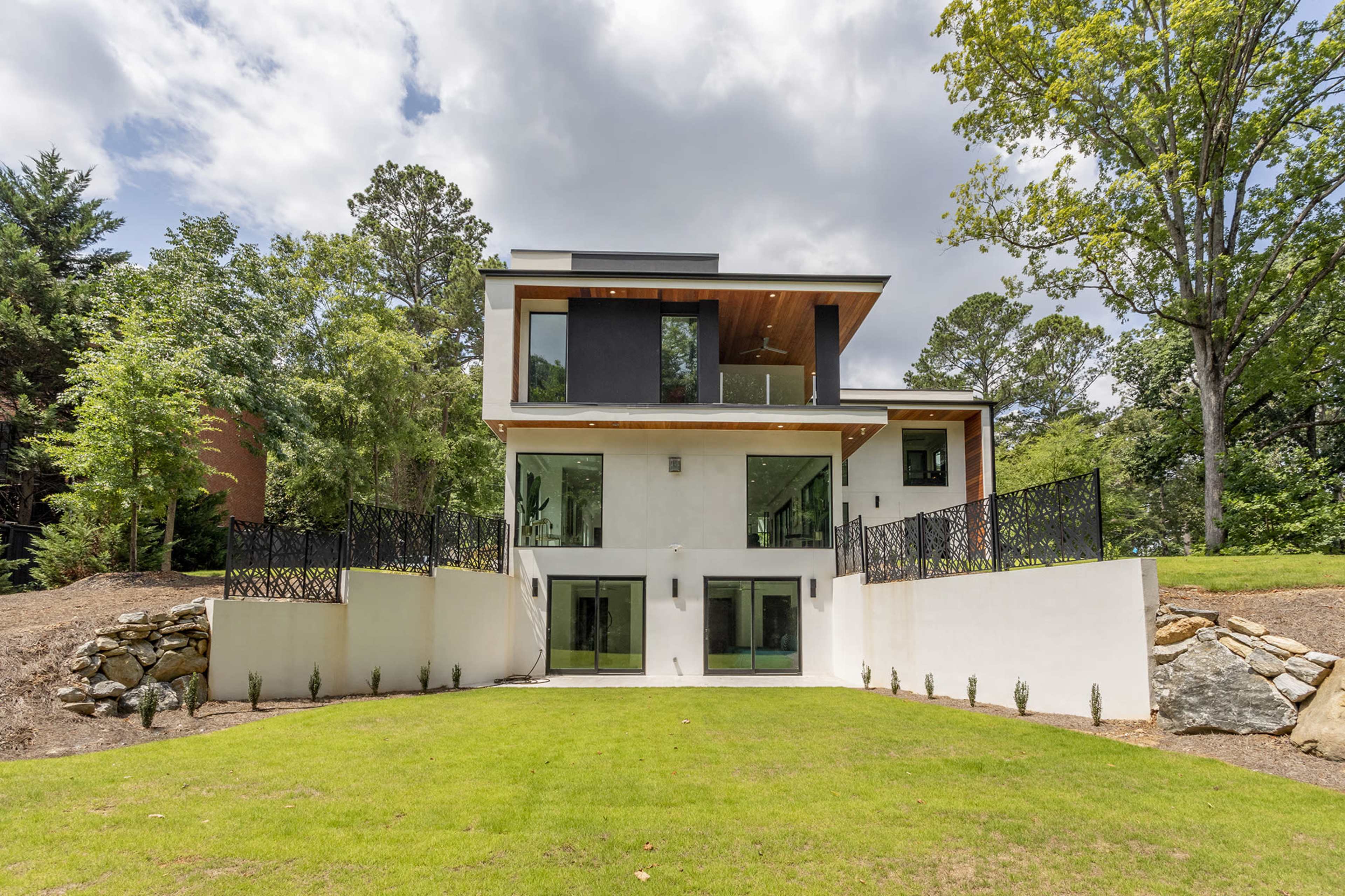 A modern three-story house with large glass windows and a grassy lawn, surrounded by trees.