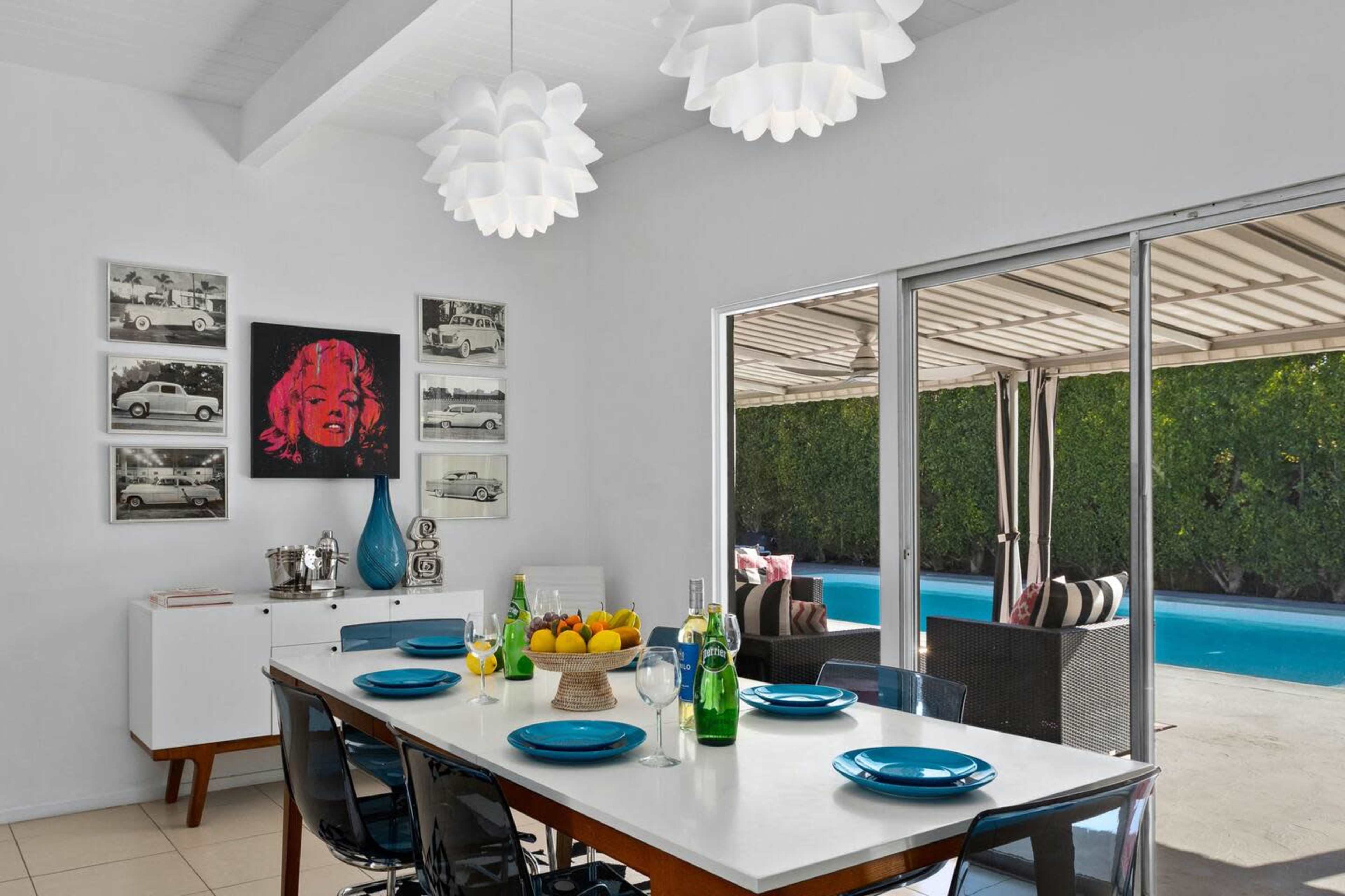 A modern dining area features a white table set with blue plates, surrounded by black chairs, while facing a large window that opens to a poolside patio.