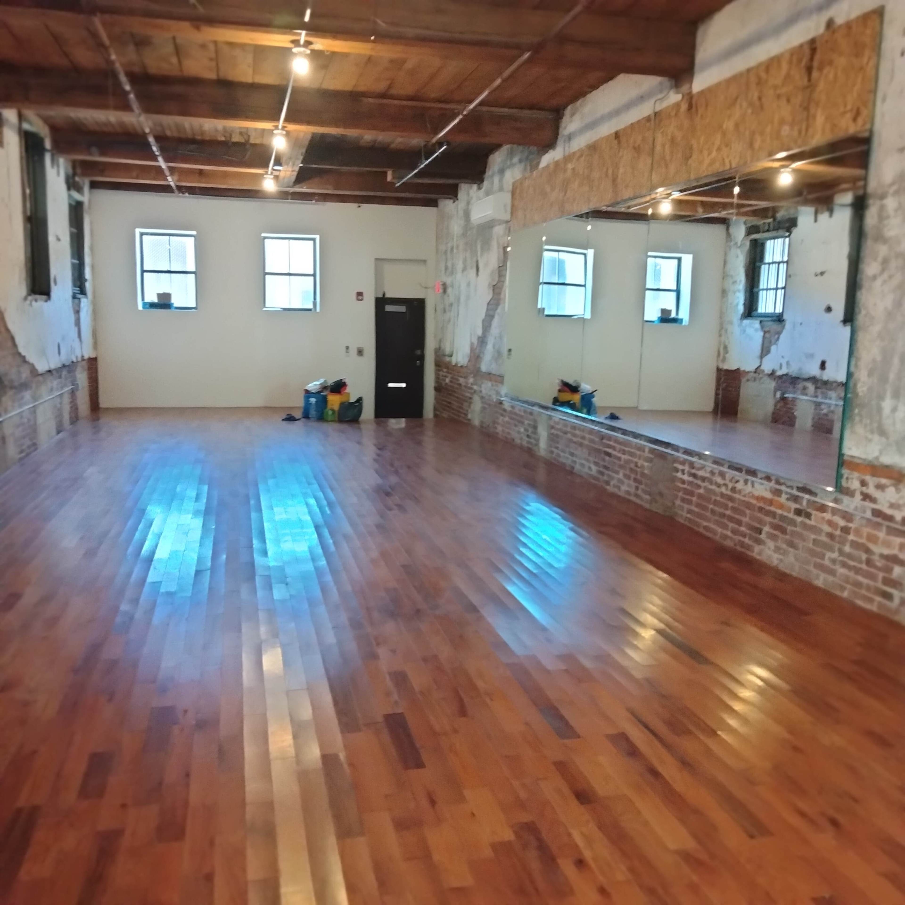 The image shows a spacious room with polished hardwood floors, exposed brick walls, and large mirrors along one side.