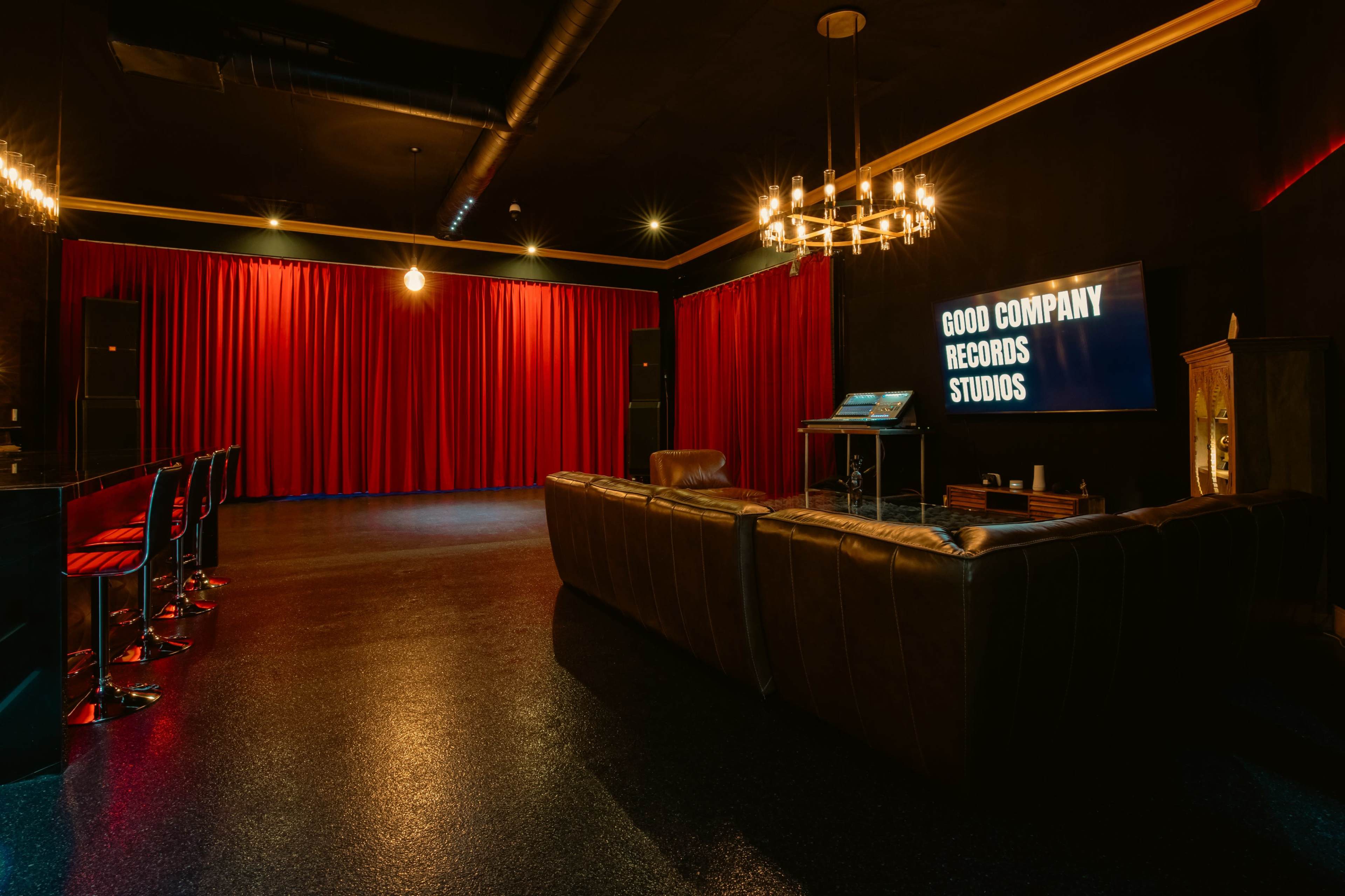 The image shows a recording studio with a dark interior, featuring a leather couch, a screen displaying "GOOD COMPANY RECORDS STUDIOS," and red curtains in the background.