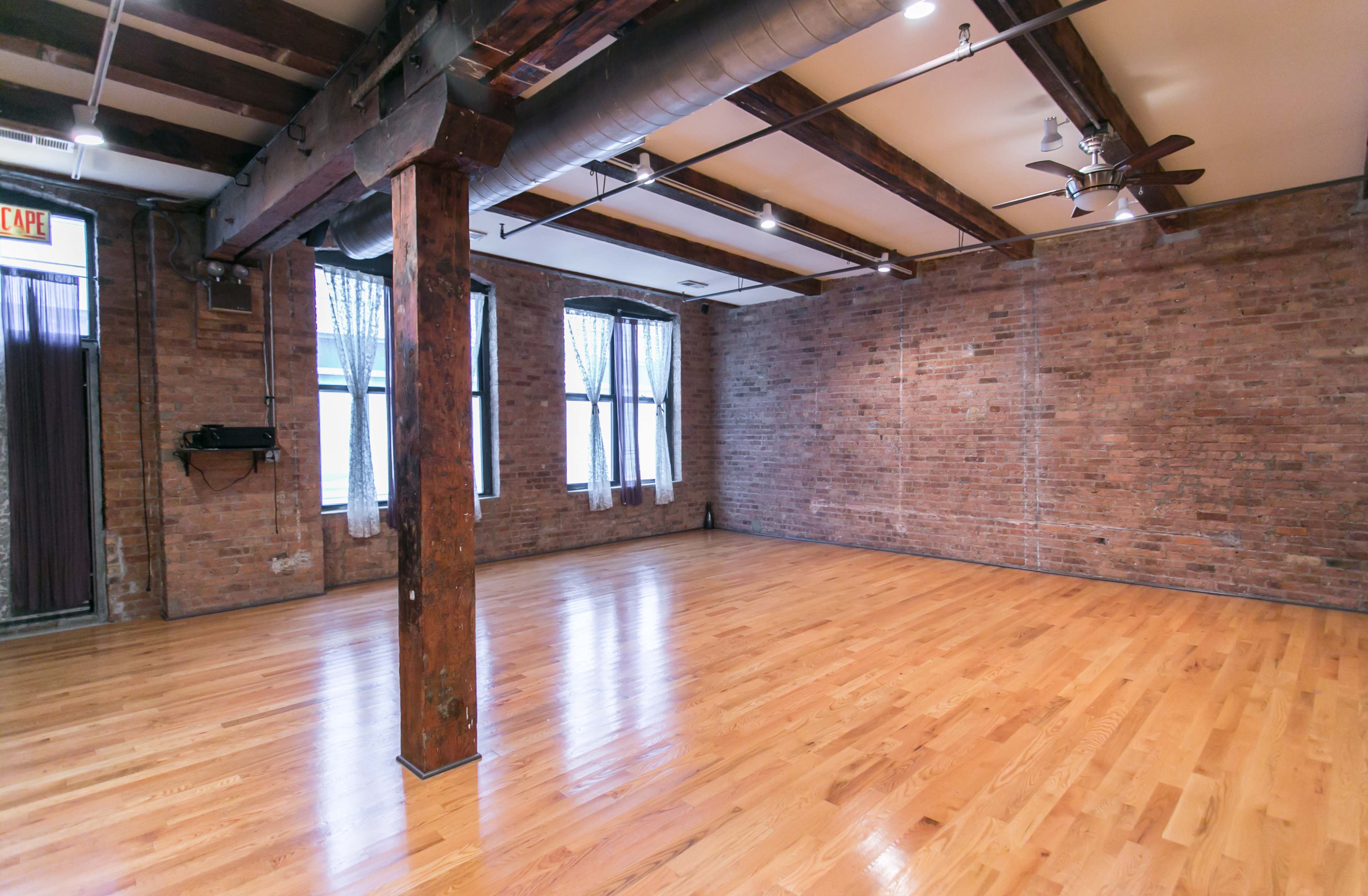 An empty room with wooden flooring, exposed brick walls, and large windows.