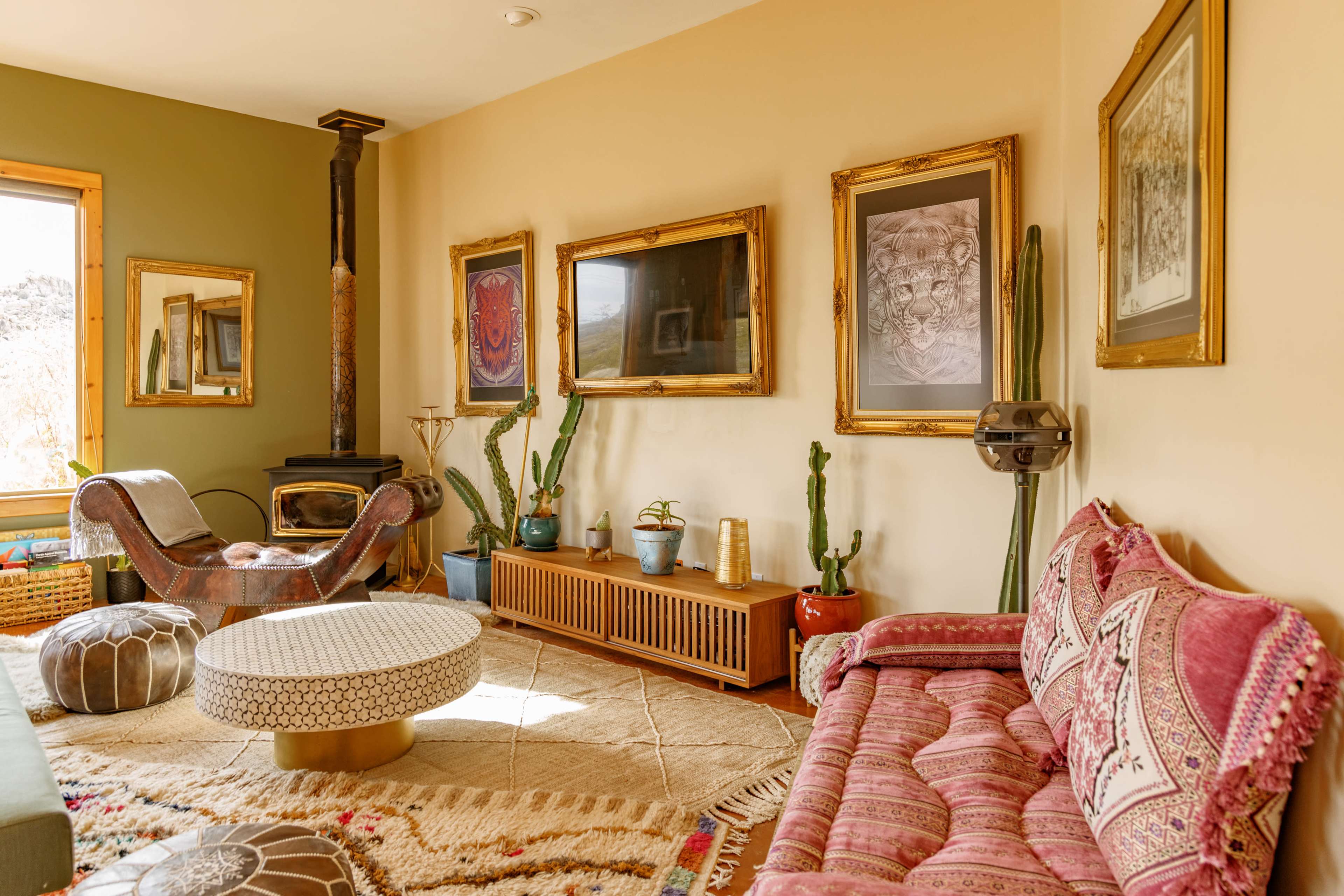 The room features a mix of vintage furniture, decorative art, and potted plants, with a fireplace in one corner and large windows allowing natural light.