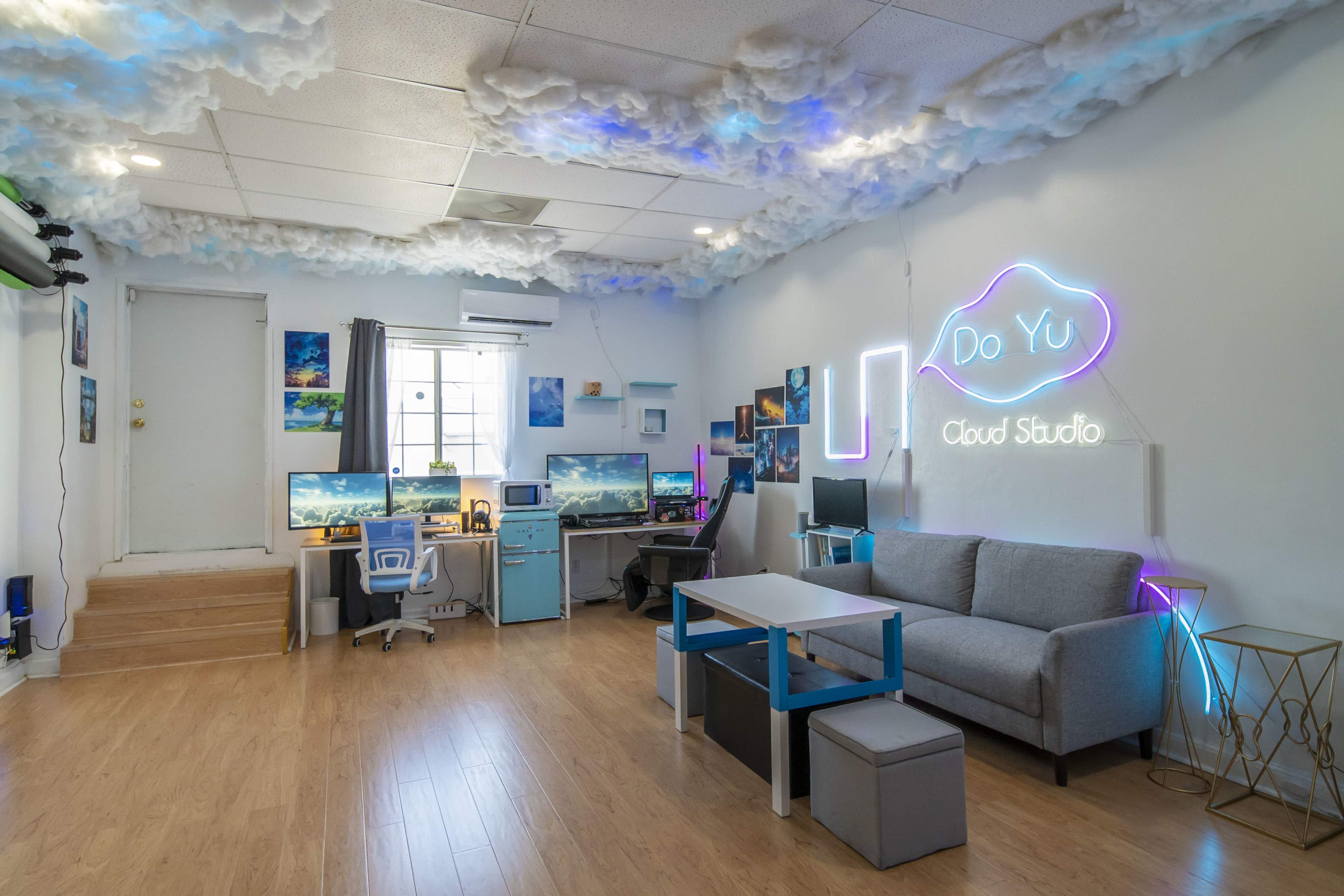 A modern cloud-themed studio featuring two computer desks, a couch, and decorative wall art, with a neon sign that says "Do Yu Cloud Studio."