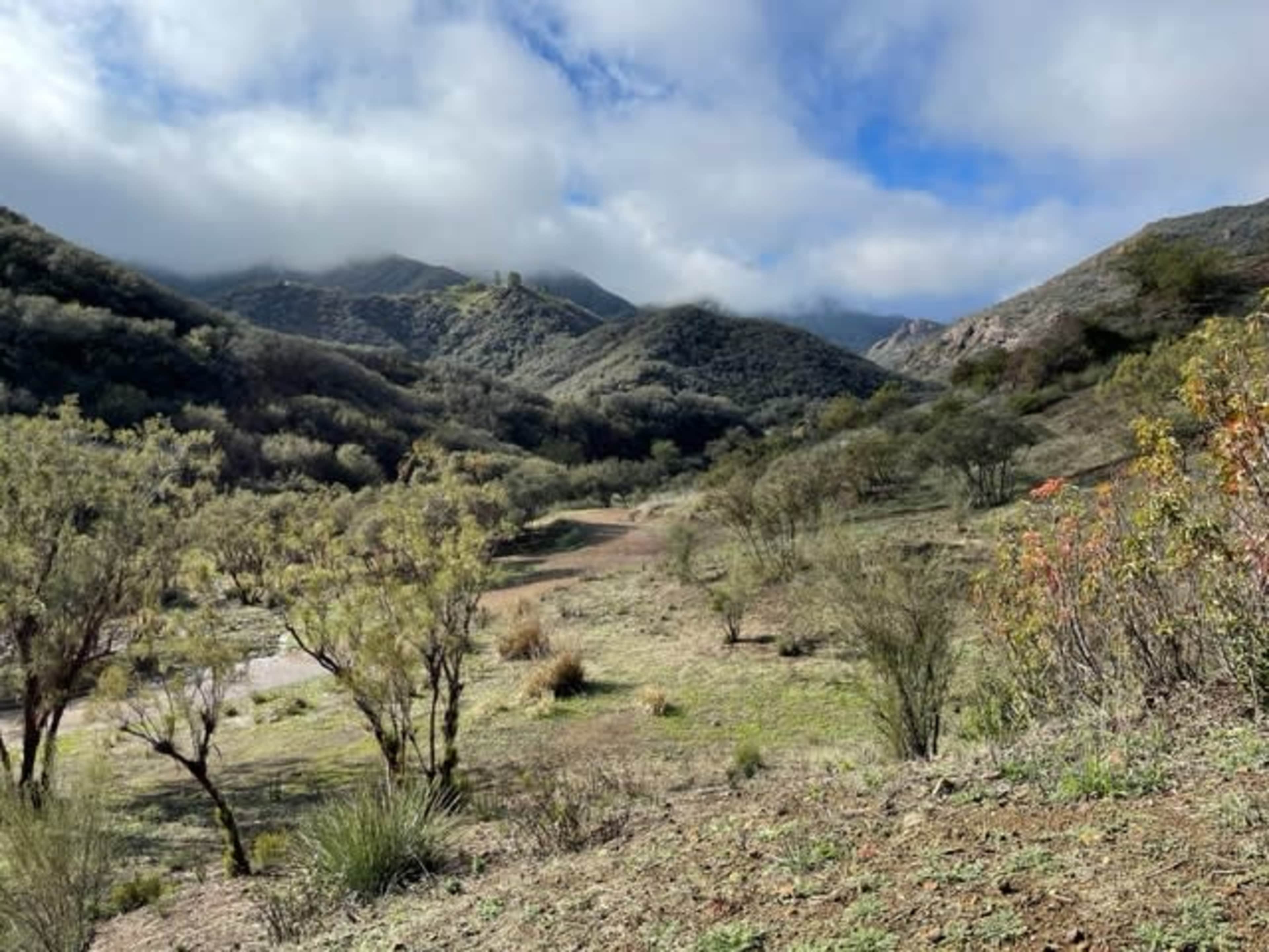 A winding dirt path leads through a hilly landscape covered with sparse vegetation under a cloudy sky.
