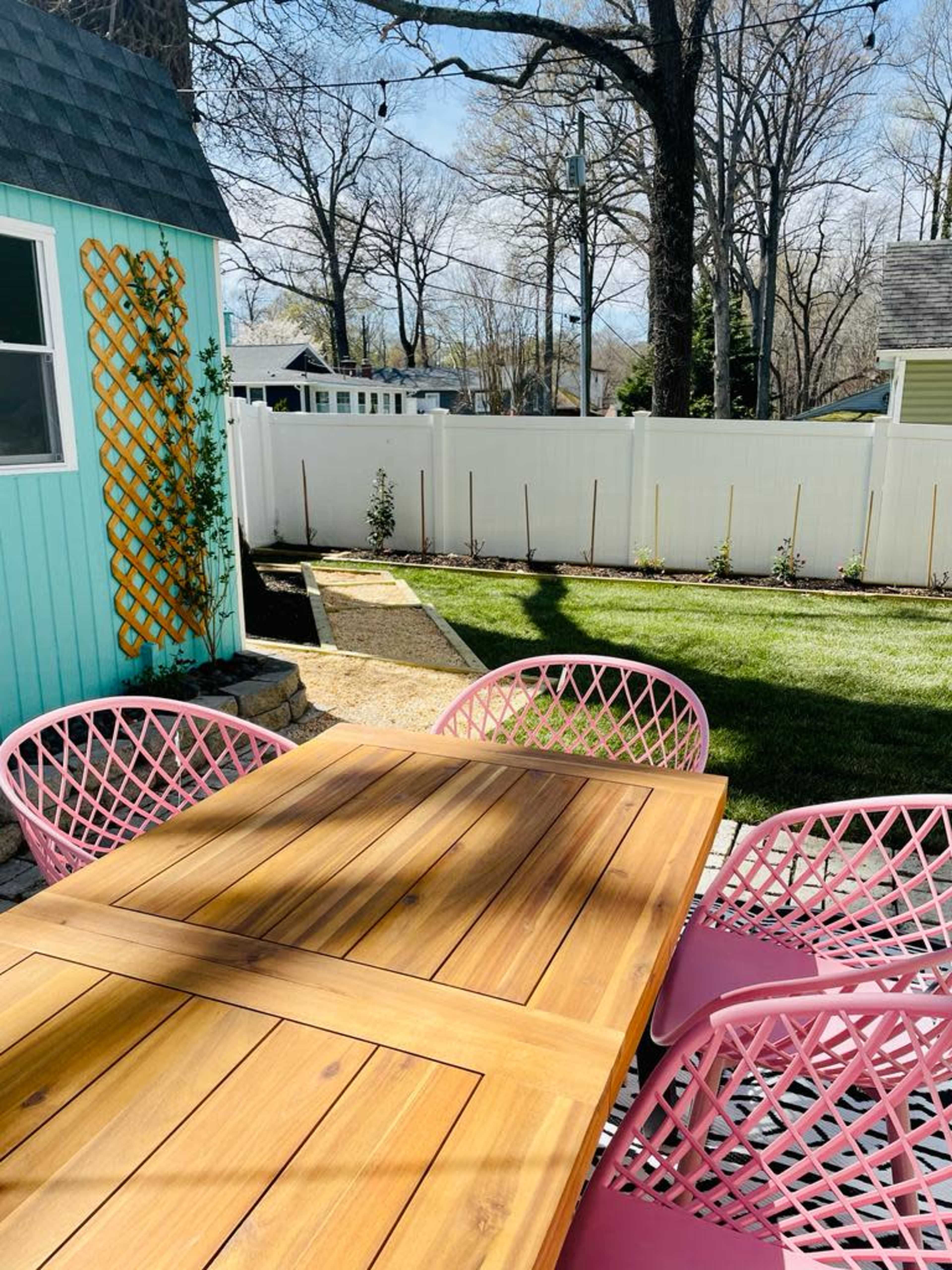 A wooden table with pink chairs is set in a backyard featuring a green lawn and a white fence.