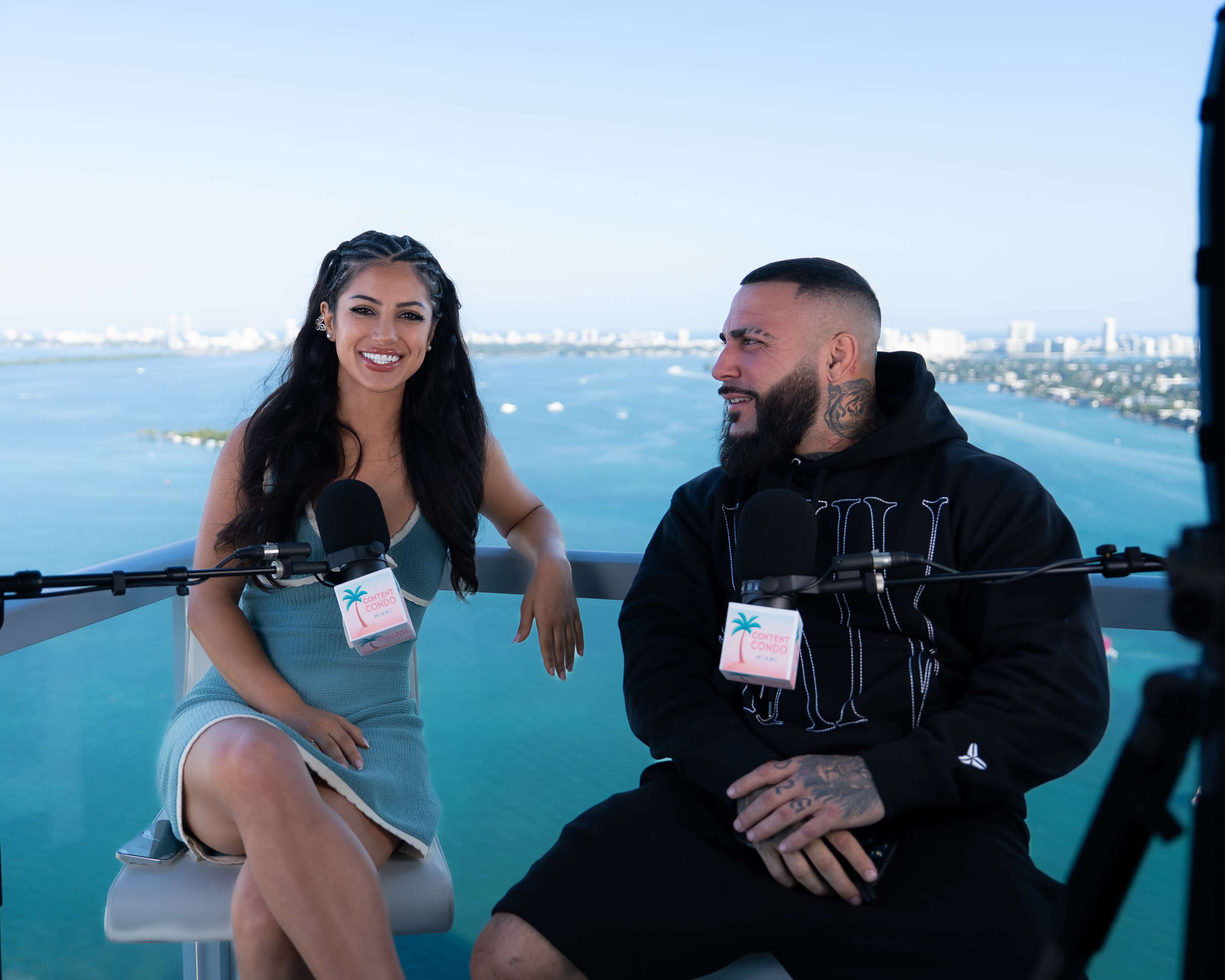 Two individuals are seated at a table with microphones, overlooking a body of water and a city skyline in the background.