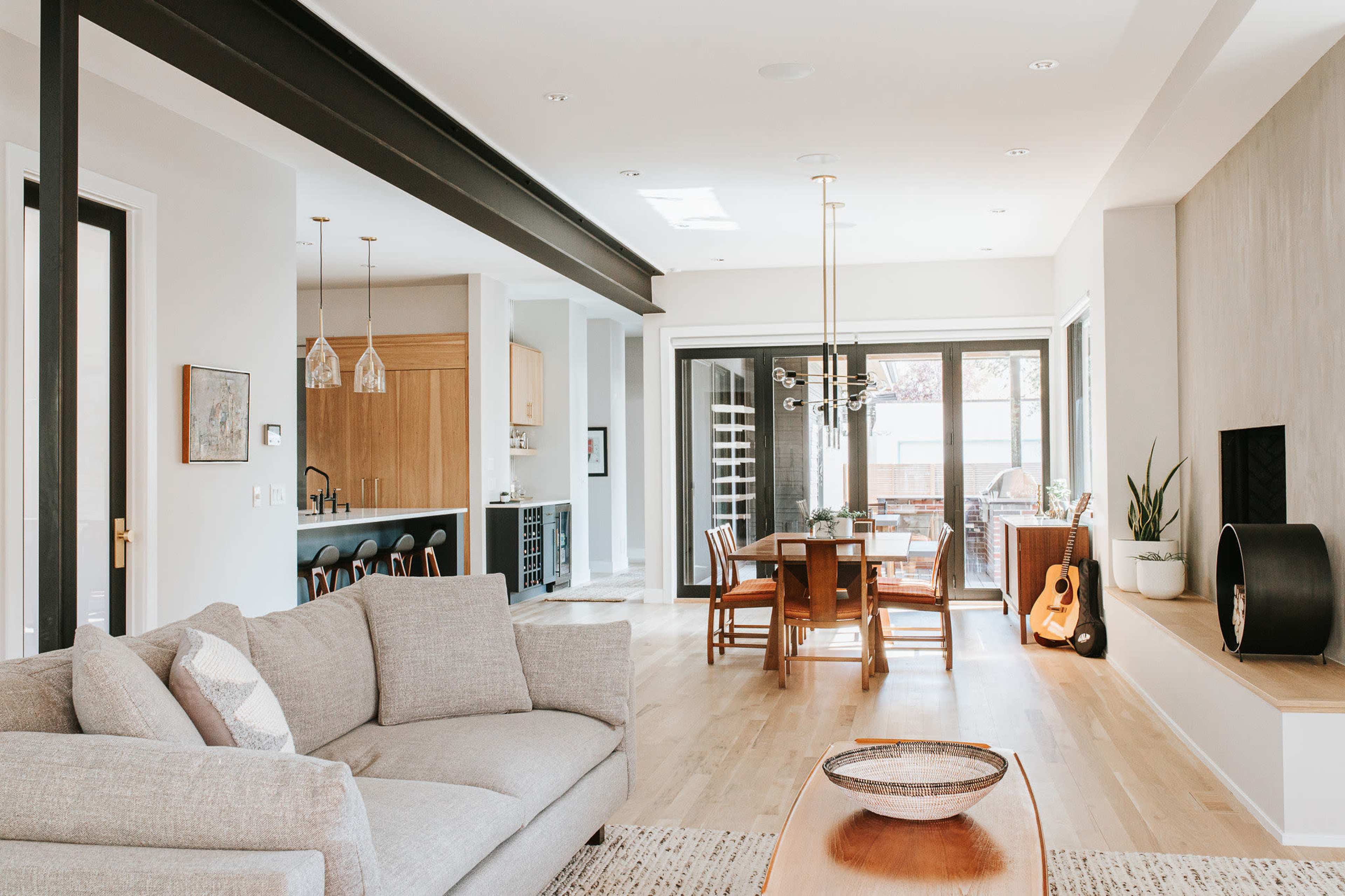 A modern open-concept living and dining area features a light-colored sofa, wooden dining table, and large windows providing ample natural light.
