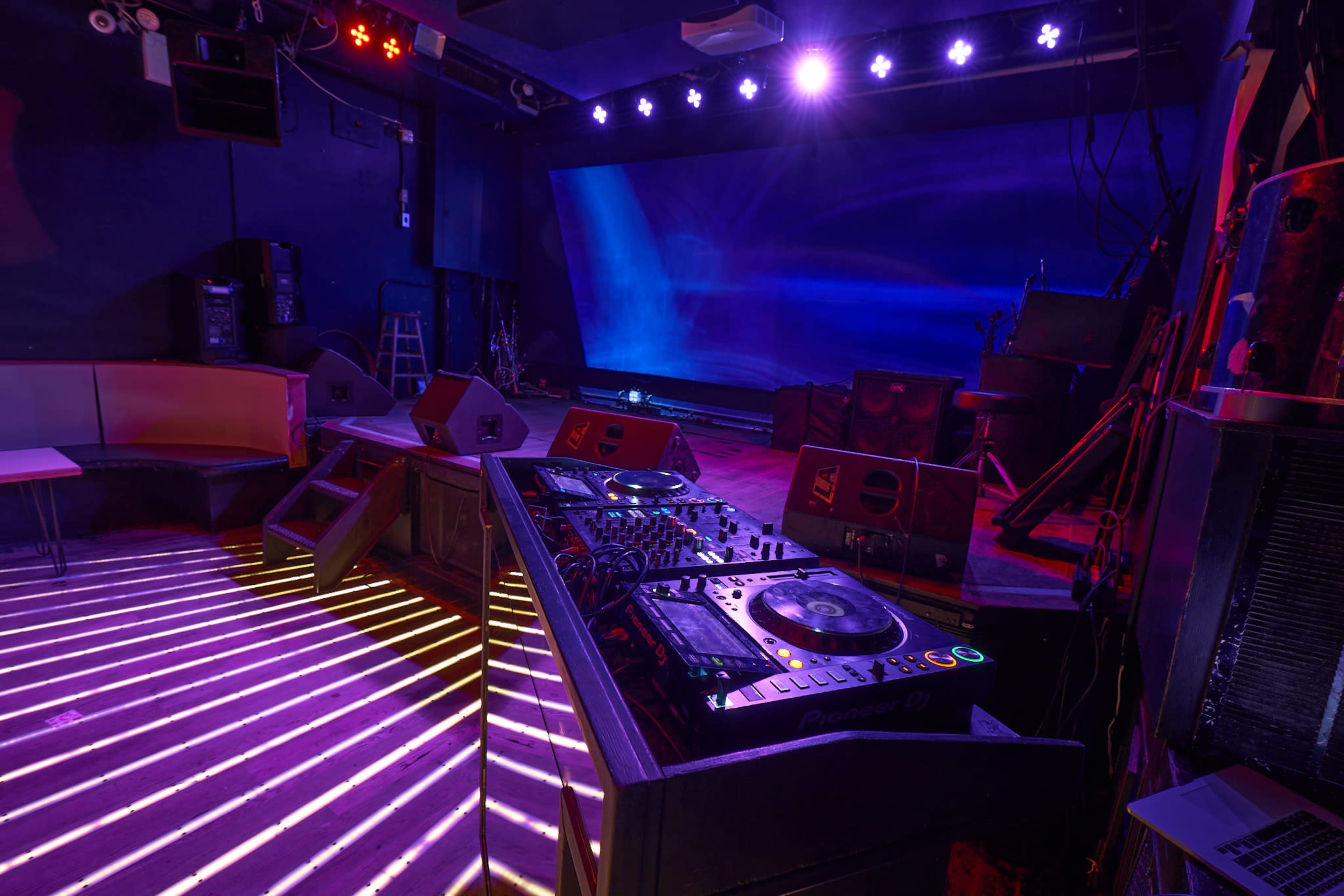 A dimly lit music venue with a DJ booth, various speakers, and a large screen in the background.