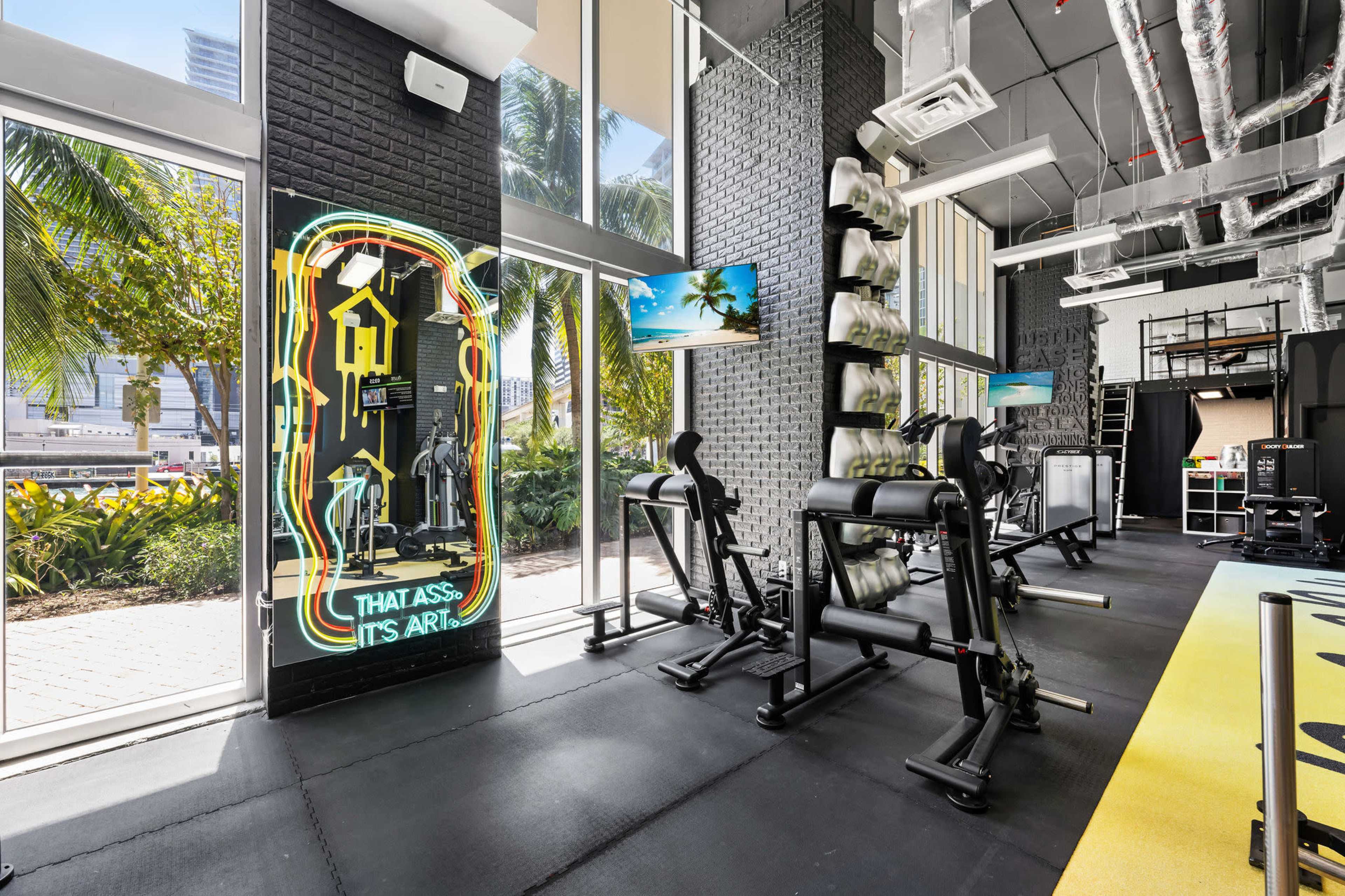 A modern gym interior with workout equipment, large windows, and vibrant decor featuring a neon sign.
