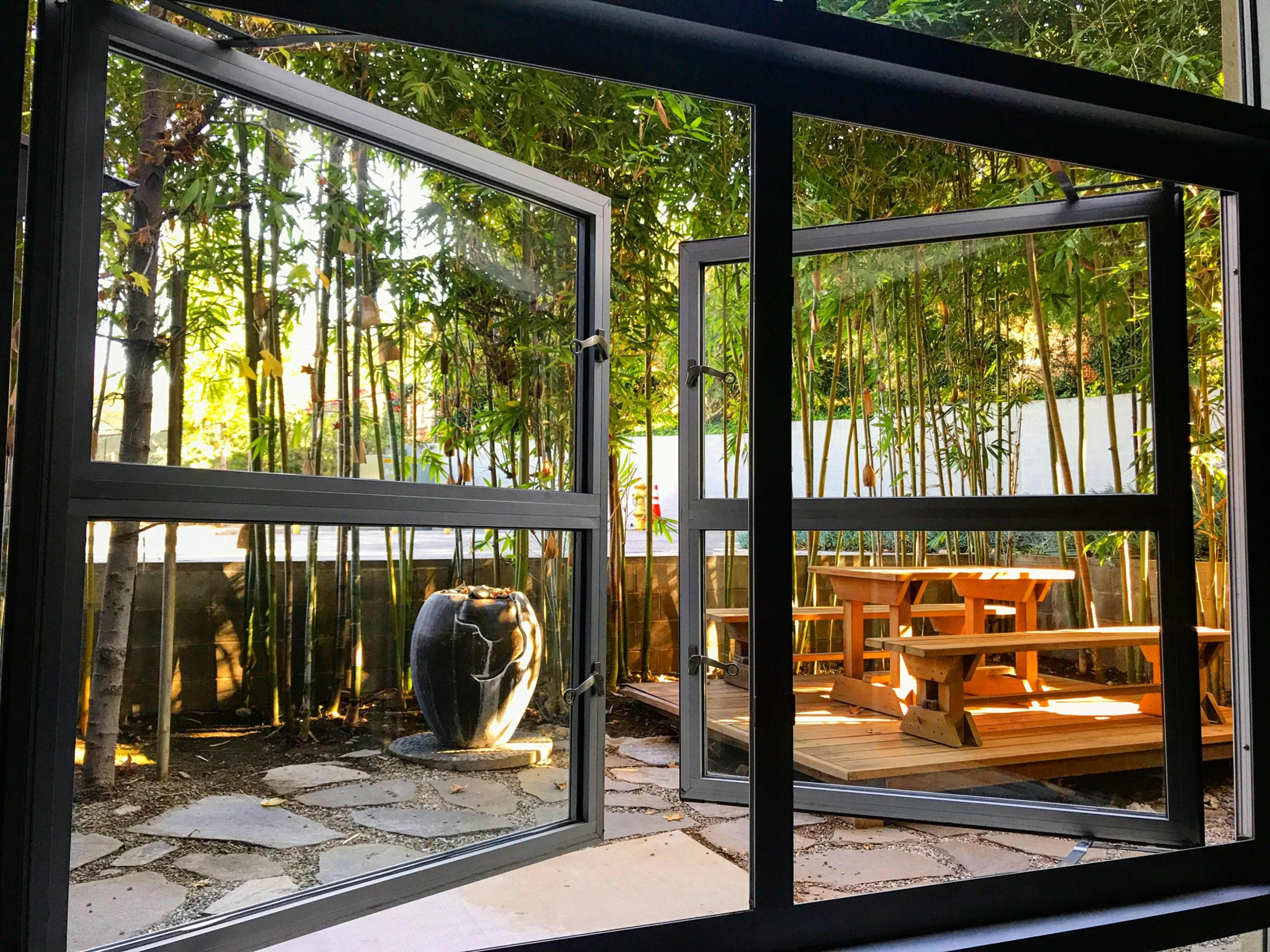 A pair of open windows frames a view of a bamboo-filled garden with a seating area and a decorative stone fountain.