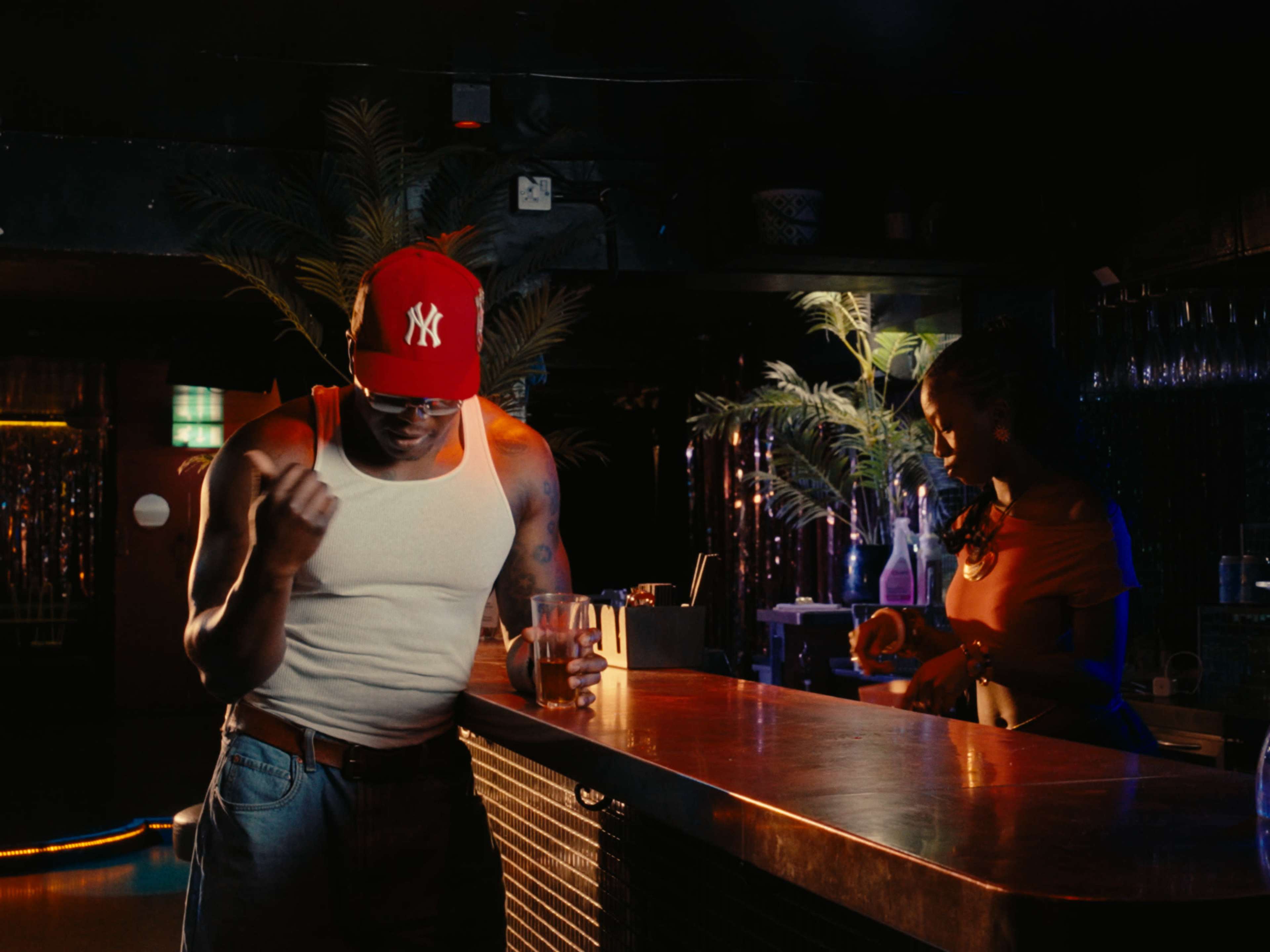 A man in a sleeveless shirt and a red baseball cap leans against a bar while a woman prepares drinks in a dimly lit nightclub.
