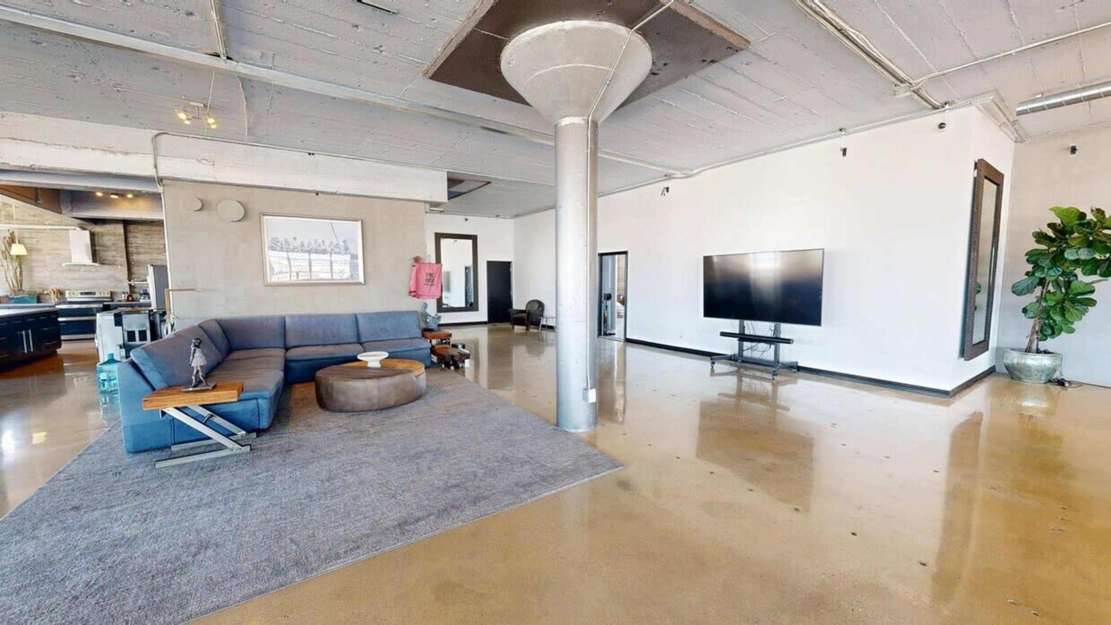 The image shows a spacious, modern loft with polished concrete flooring, a large sectional sofa, a television stand, and a decorative plant.