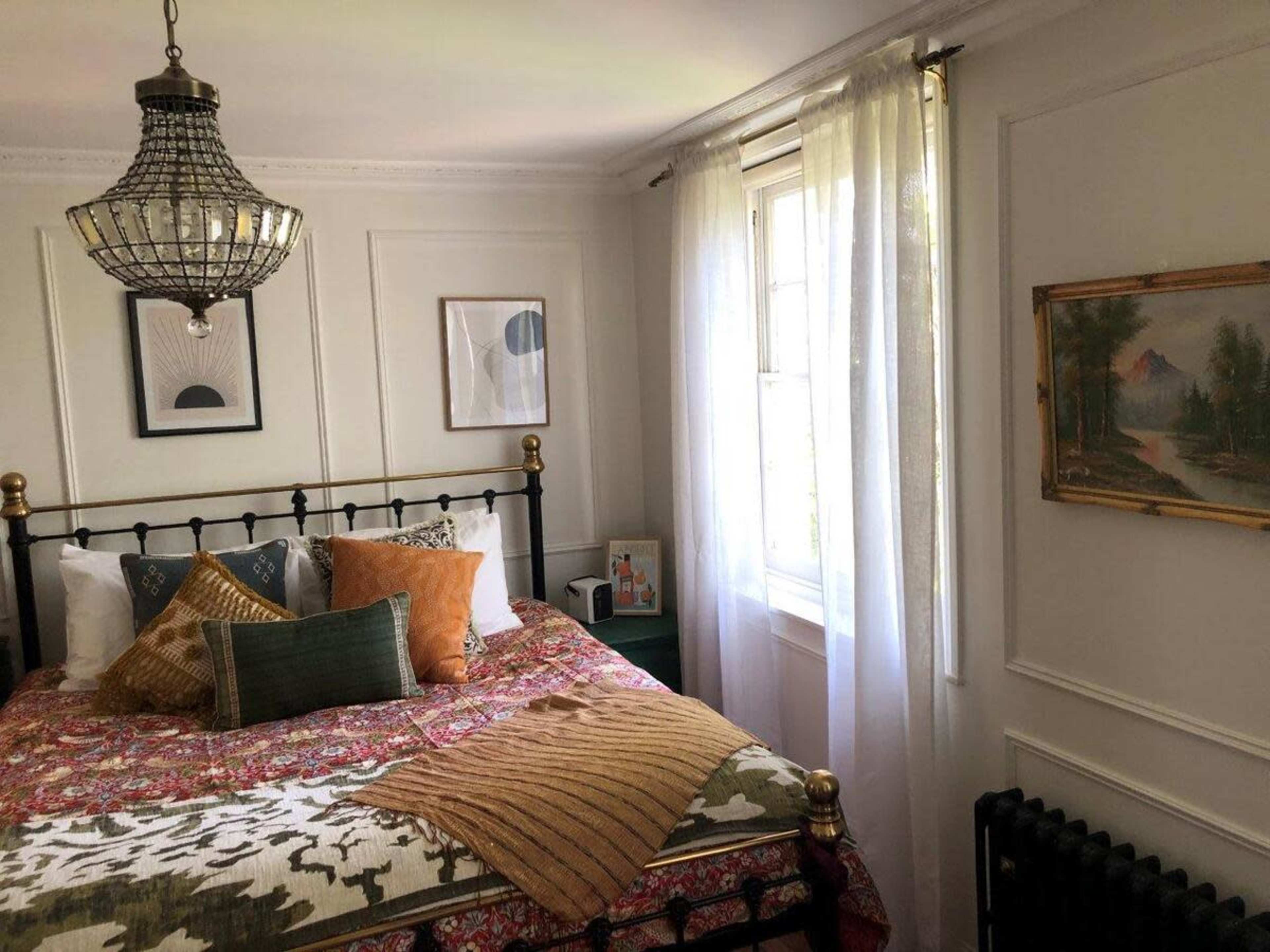 A neatly arranged bedroom featuring a wrought iron bed with colorful bedding, a large window with sheer curtains, and a decorative ceiling light fixture.