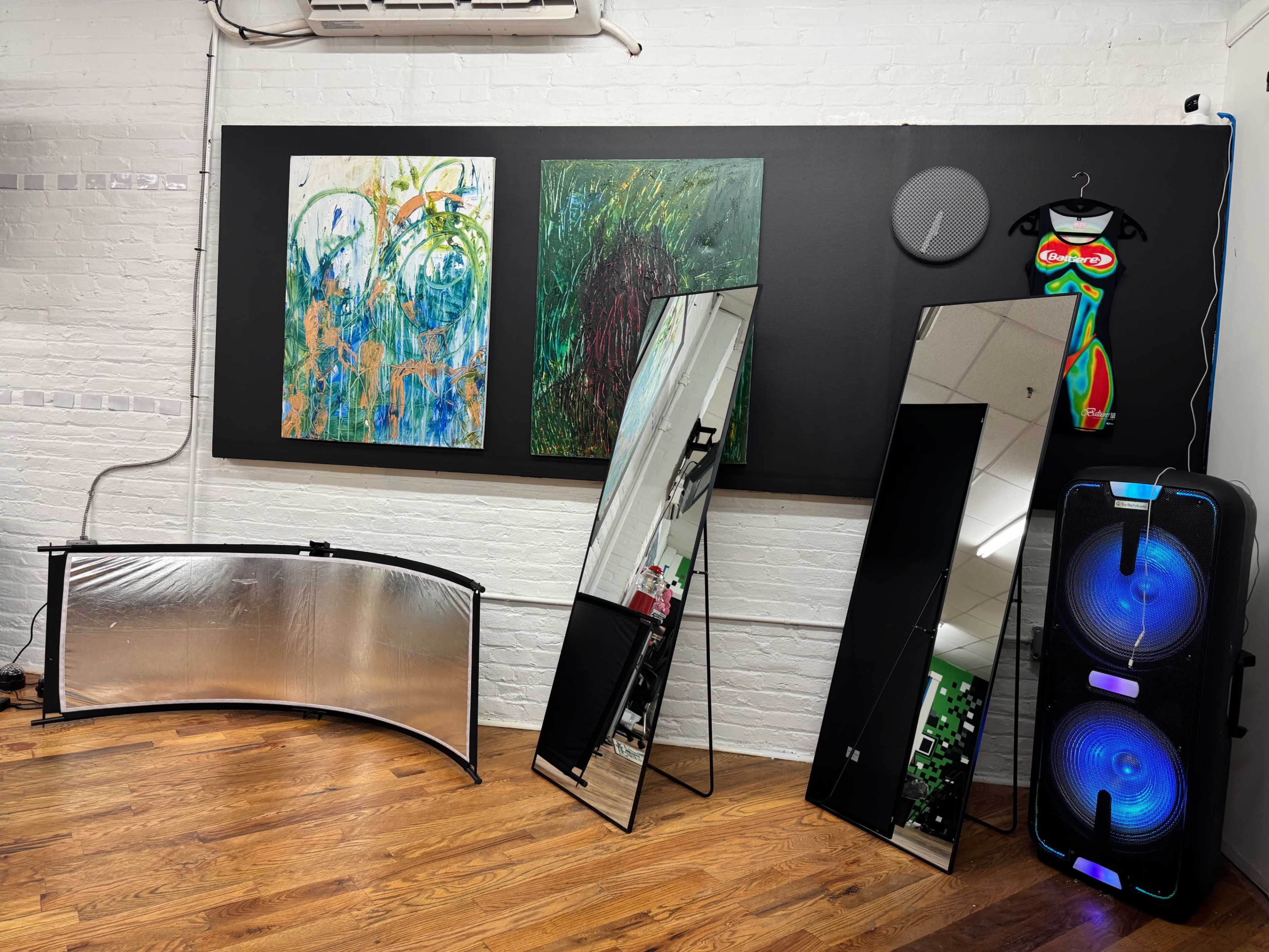 The image shows a gallery interior featuring two colorful abstract paintings on a black wall, three standing mirrors, a curved reflector, and a speaker with illuminated accents against a wooden floor.