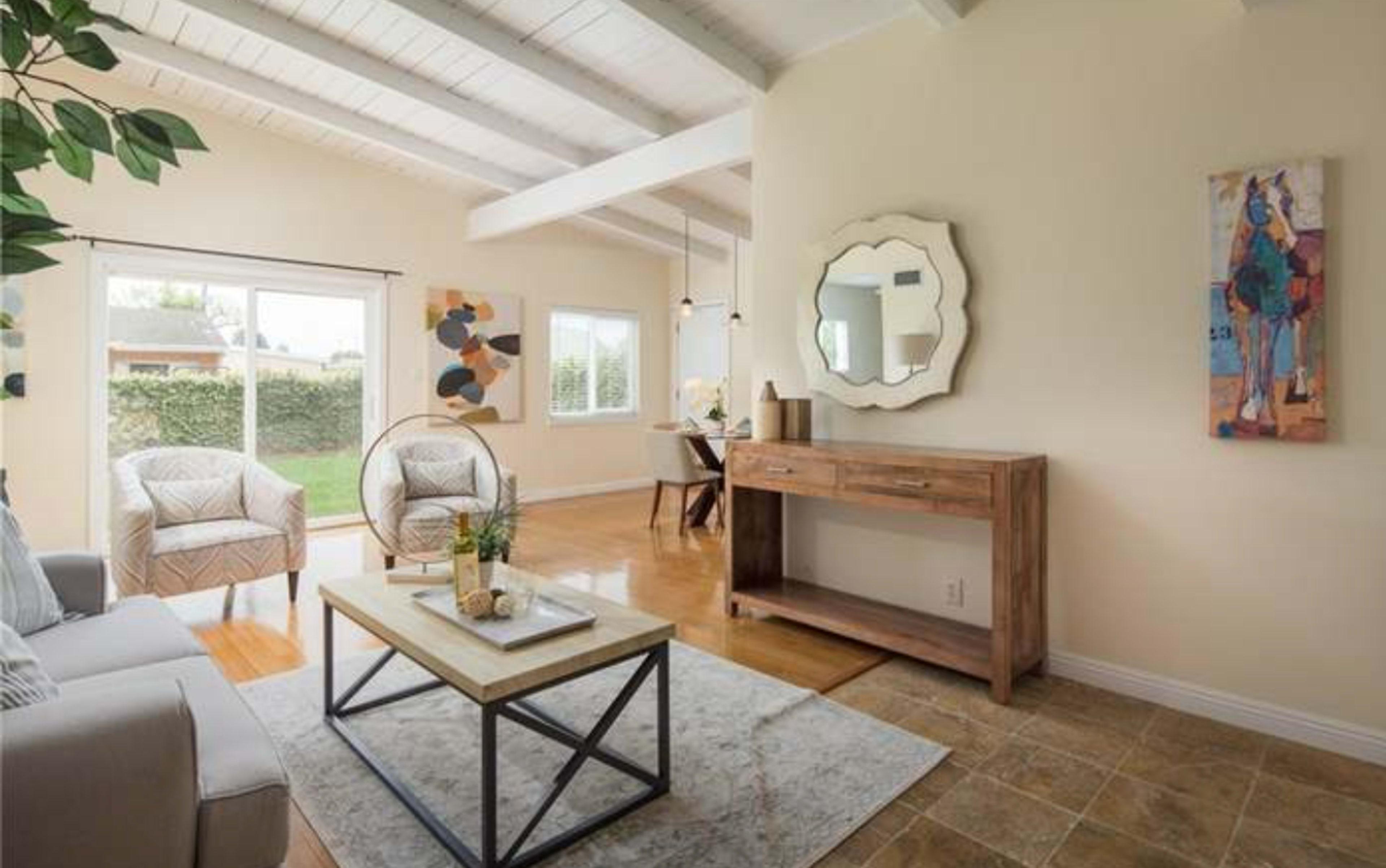 A bright living room with a light-colored sofa, a wooden coffee table on a carpet, and a console table against the wall, featuring artwork and large windows that overlook a garden.