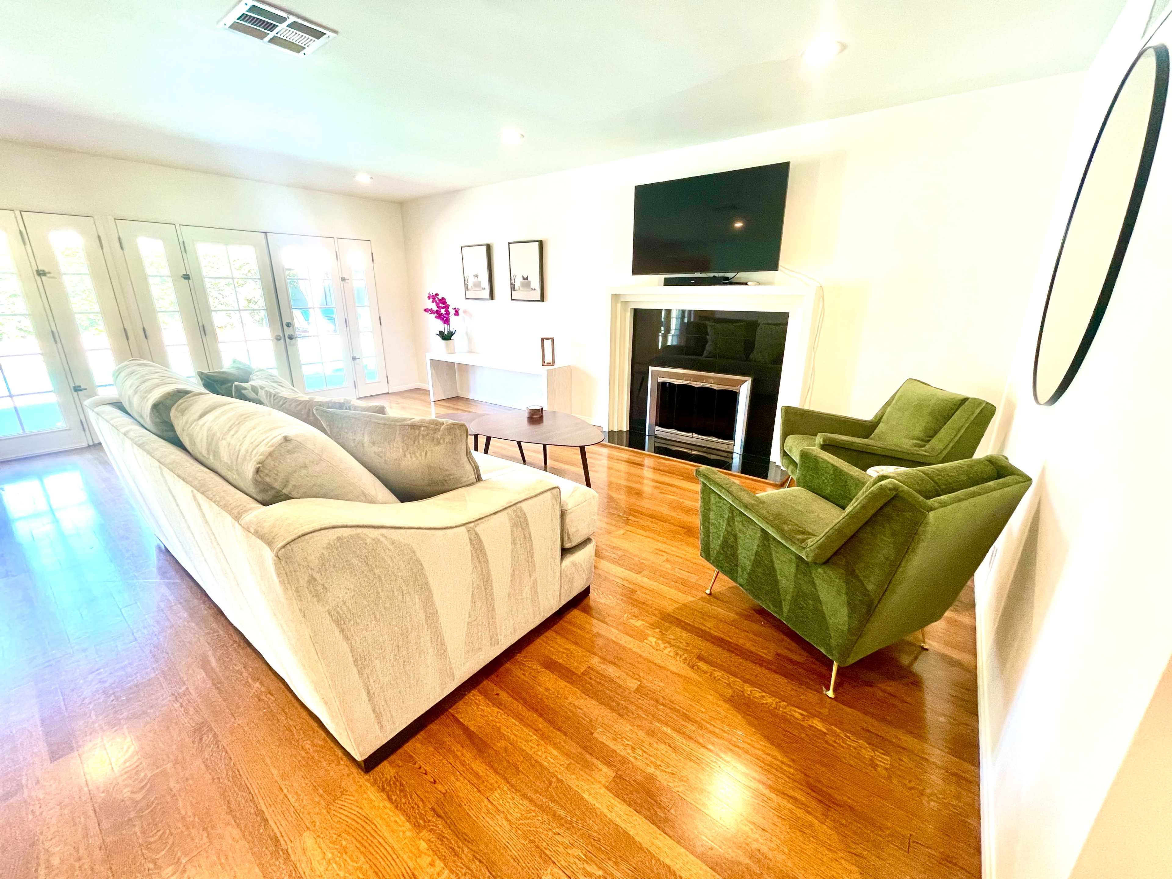 A living room with a light-colored sofa, two green armchairs, a wooden table, and a fireplace, with large windows in the background.