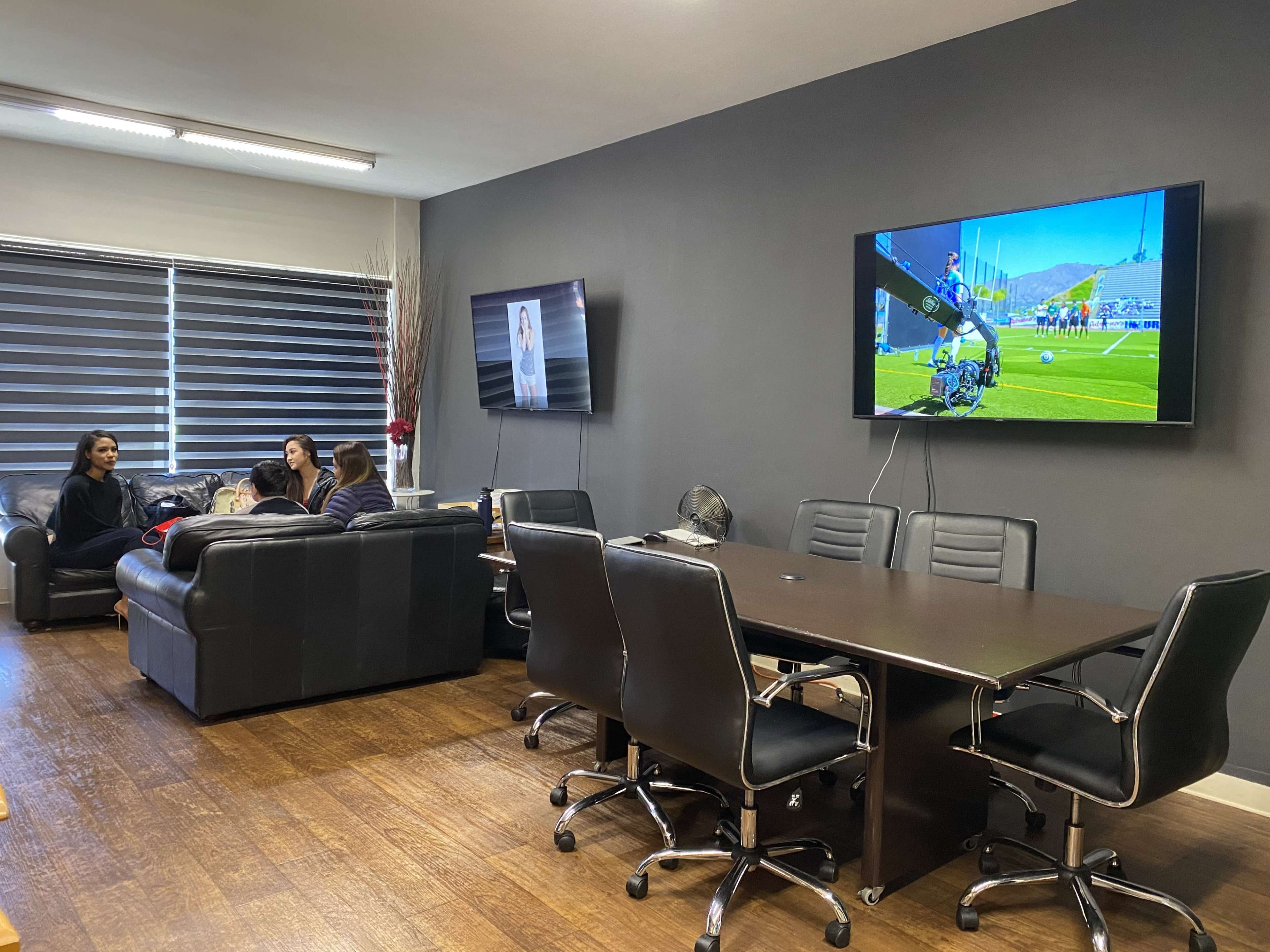 The image shows a modern office meeting room with a large table and black chairs, where three people are seated on a couch, and two screens display images on the walls.