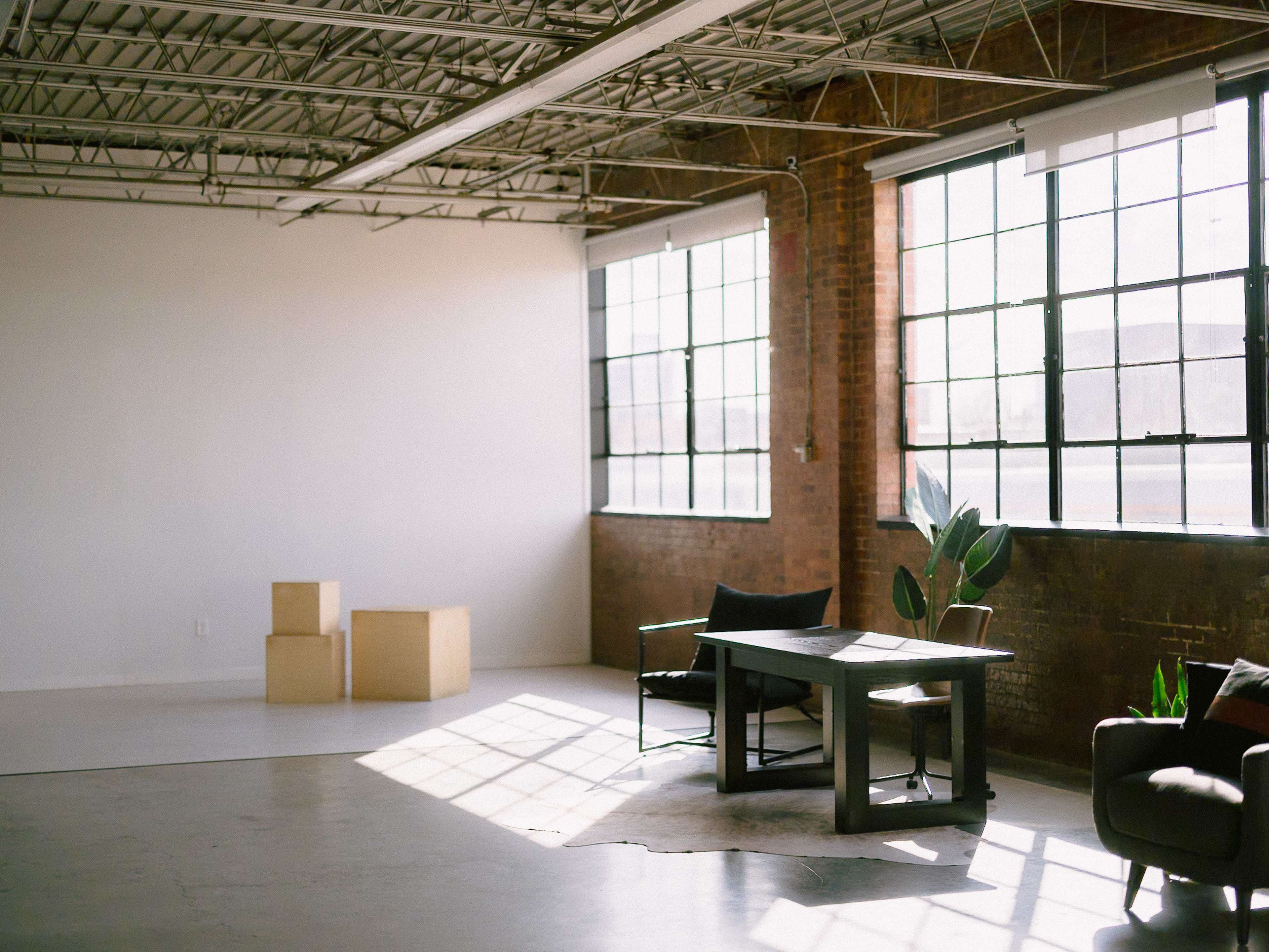 The image shows a spacious, well-lit interior with large windows, a wooden table, a chair, two cardboard boxes, and a potted plant.