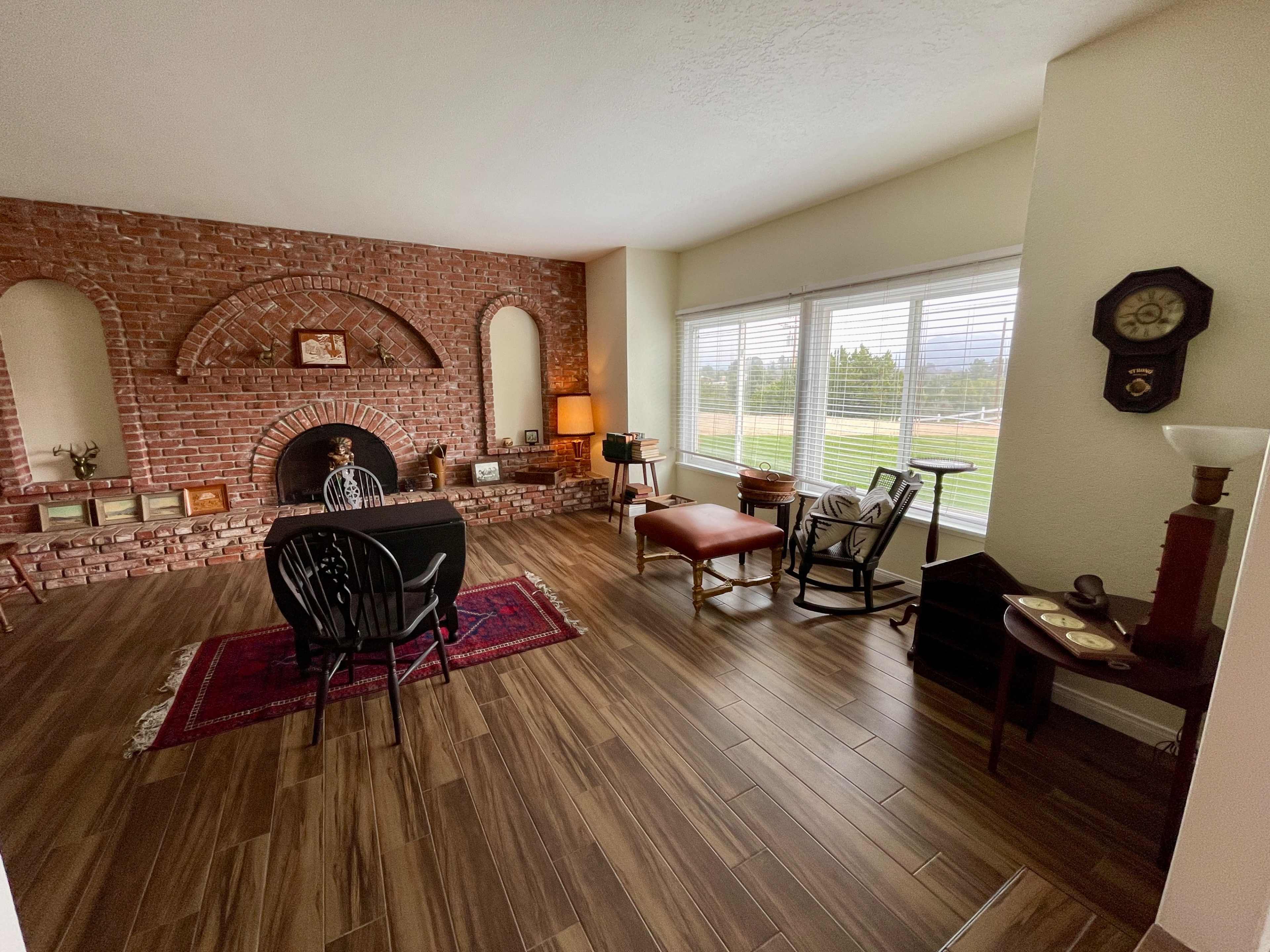 The room features a brick fireplace, a wooden table with chairs, and large windows overlooking a green landscape.