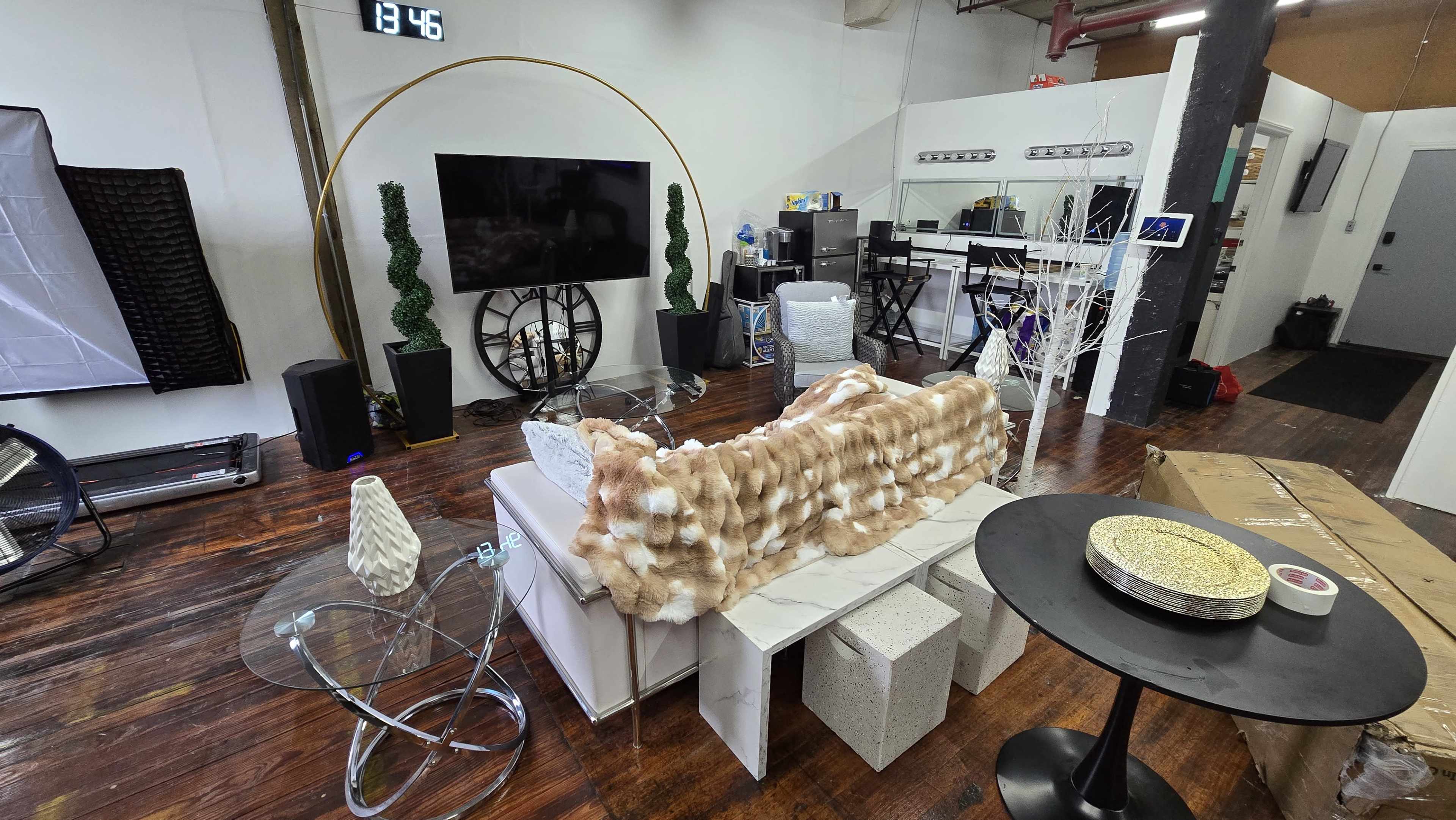 A modern interior space features a white couch with a fuzzy blanket, a glass coffee table, decorative plants, and a large television mounted on the wall.