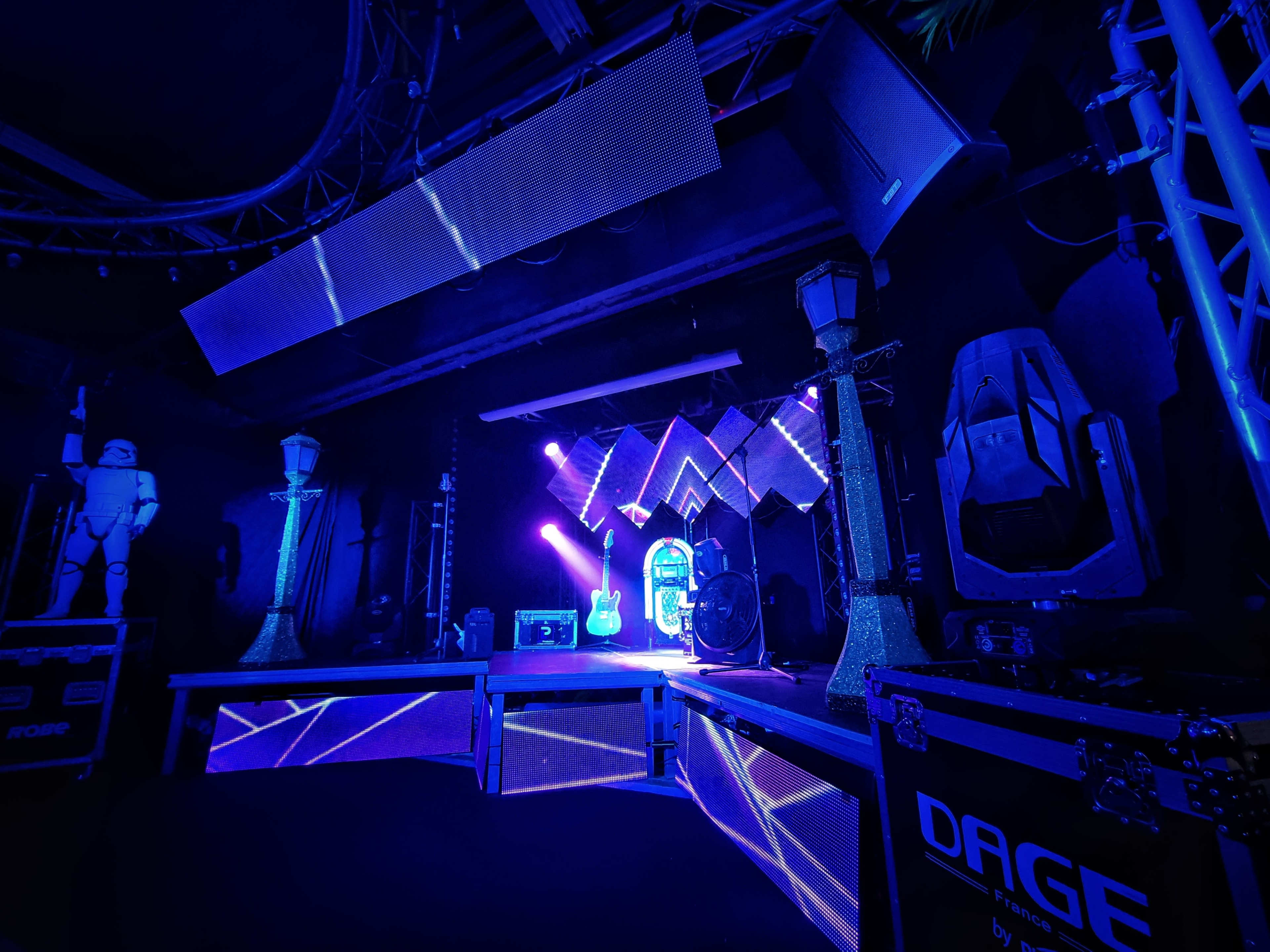 A stage setup featuring blue lighting, musical instruments, and various stage equipment in a darkened venue.