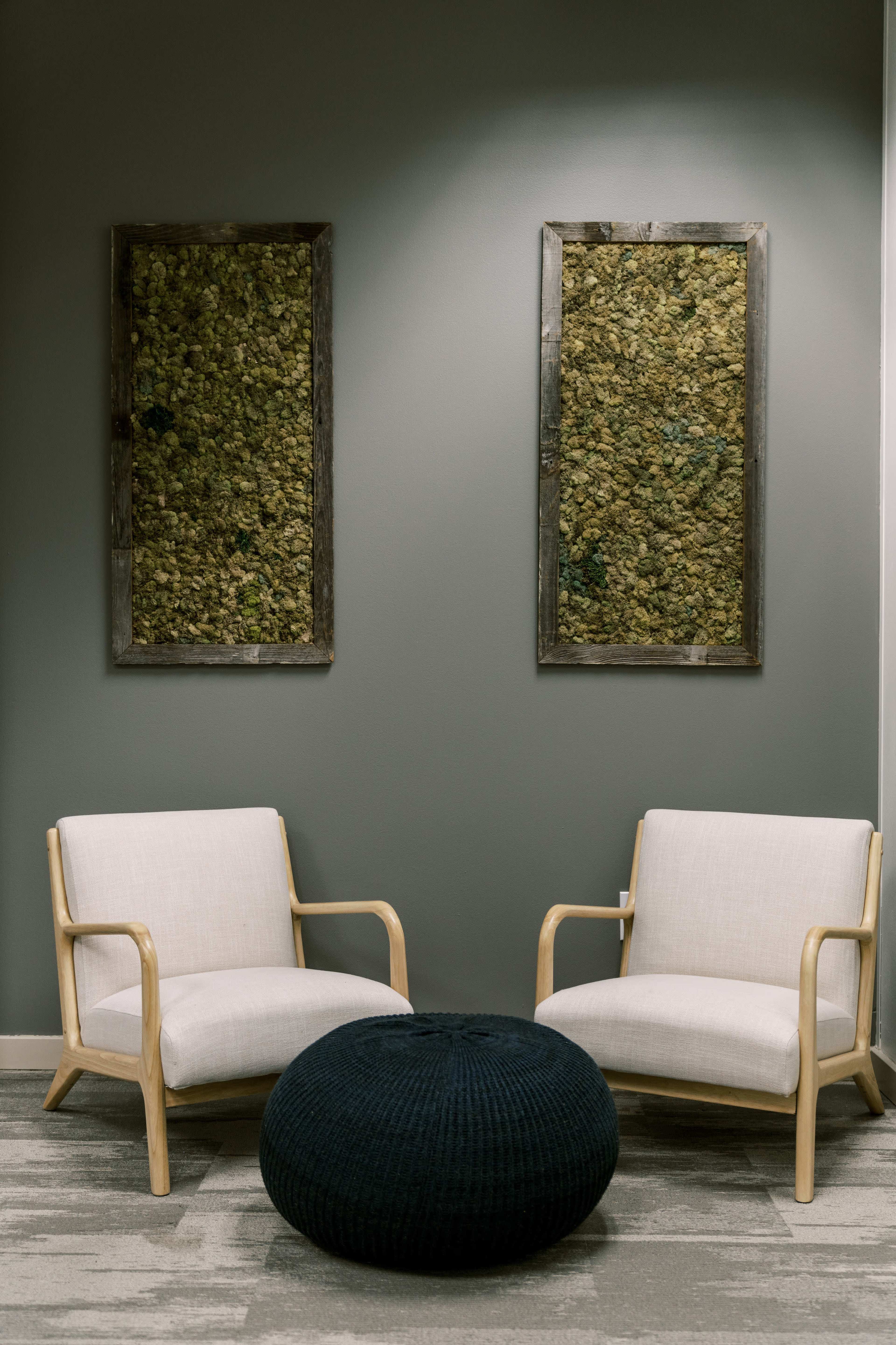 Two minimalist armchairs are positioned facing each other with a round knitted ottoman in between, set against a green wall adorned with textured art pieces.