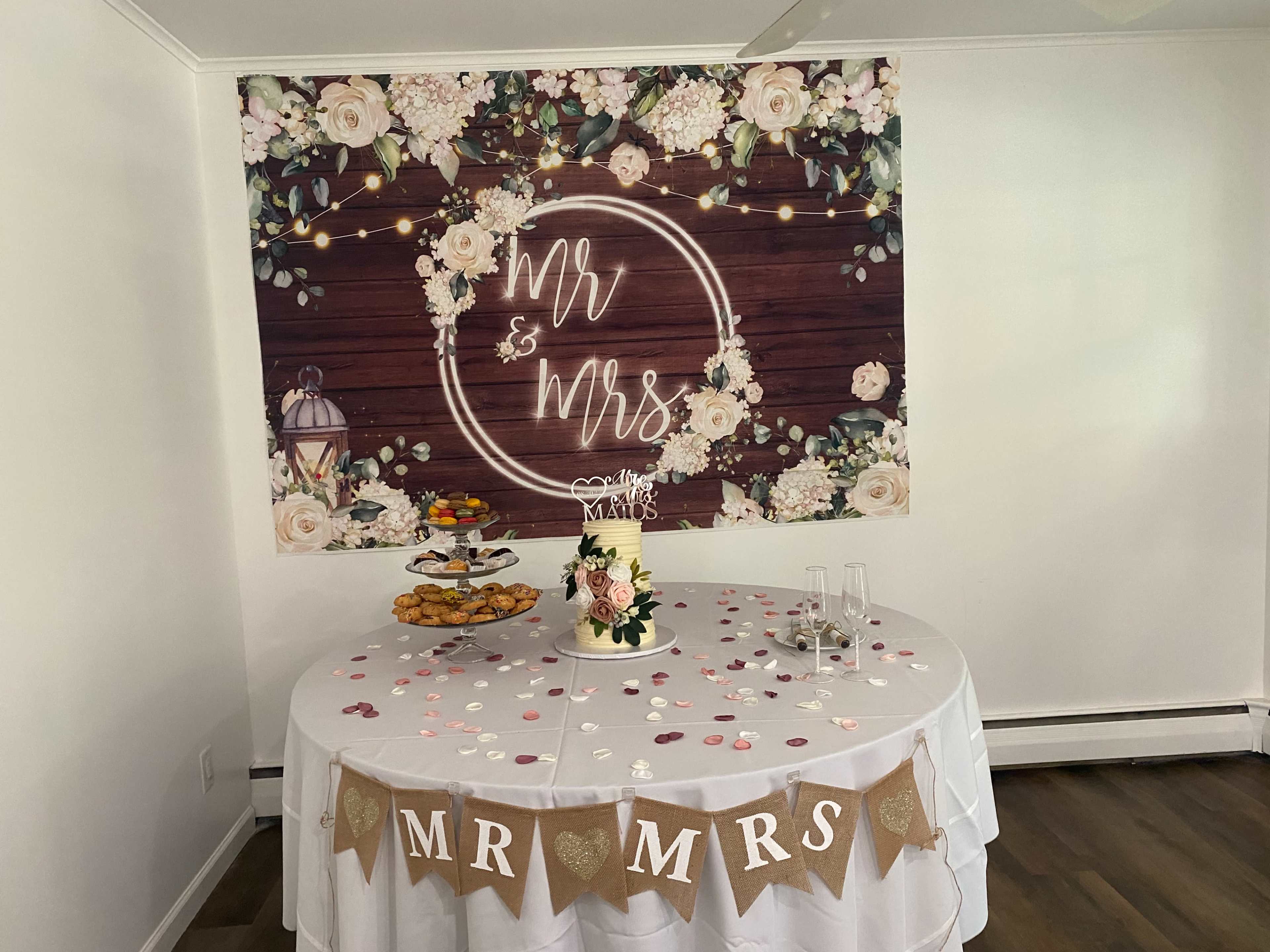 A round table decorated with a floral cake, cookies, and a banner reading "MR & MRS" is set against a backdrop featuring the same words surrounded by lights and flowers.