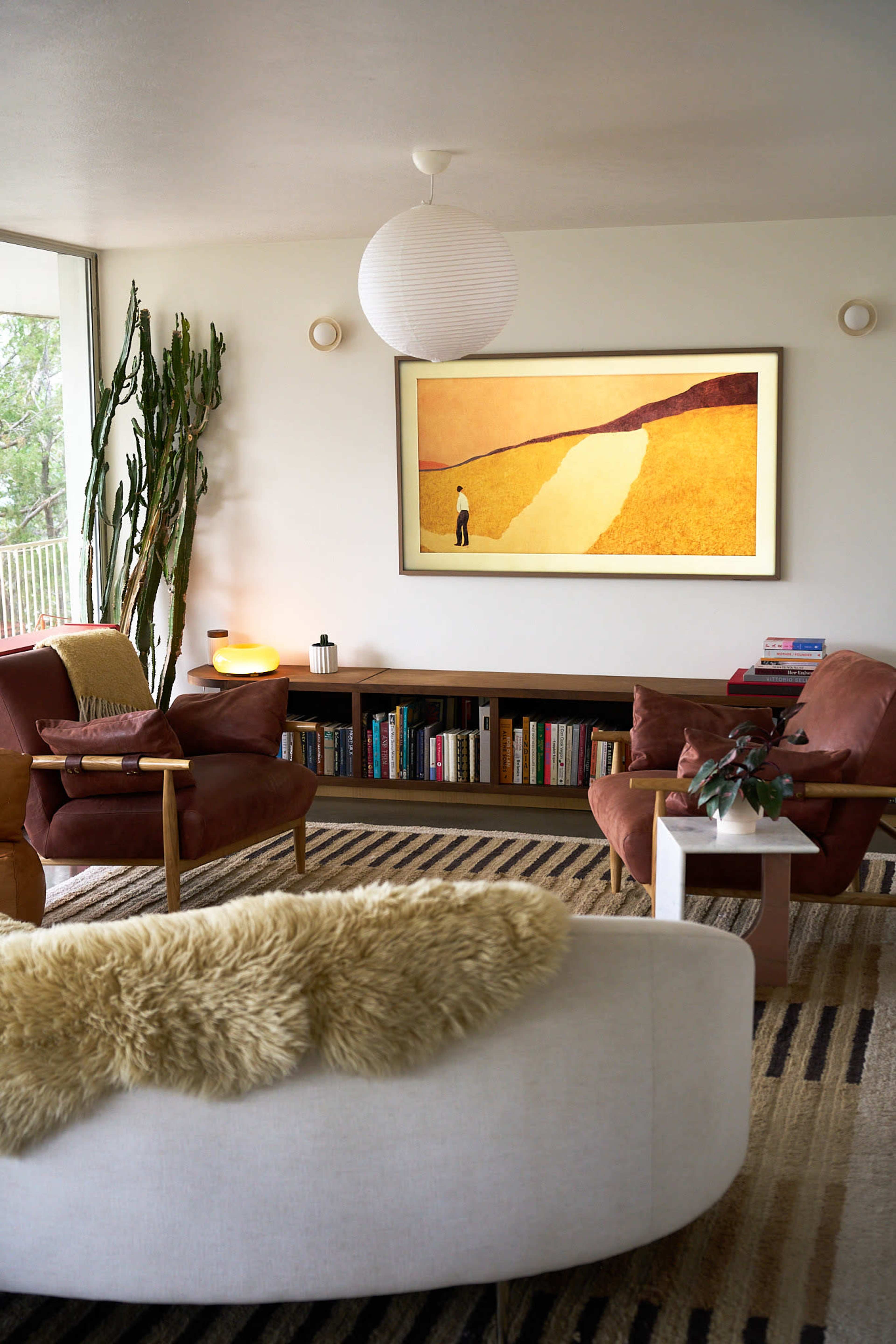 The image shows a modern living room with a sectional sofa, two armchairs, a bookshelf, and a large artwork featuring a figure in a natural landscape.