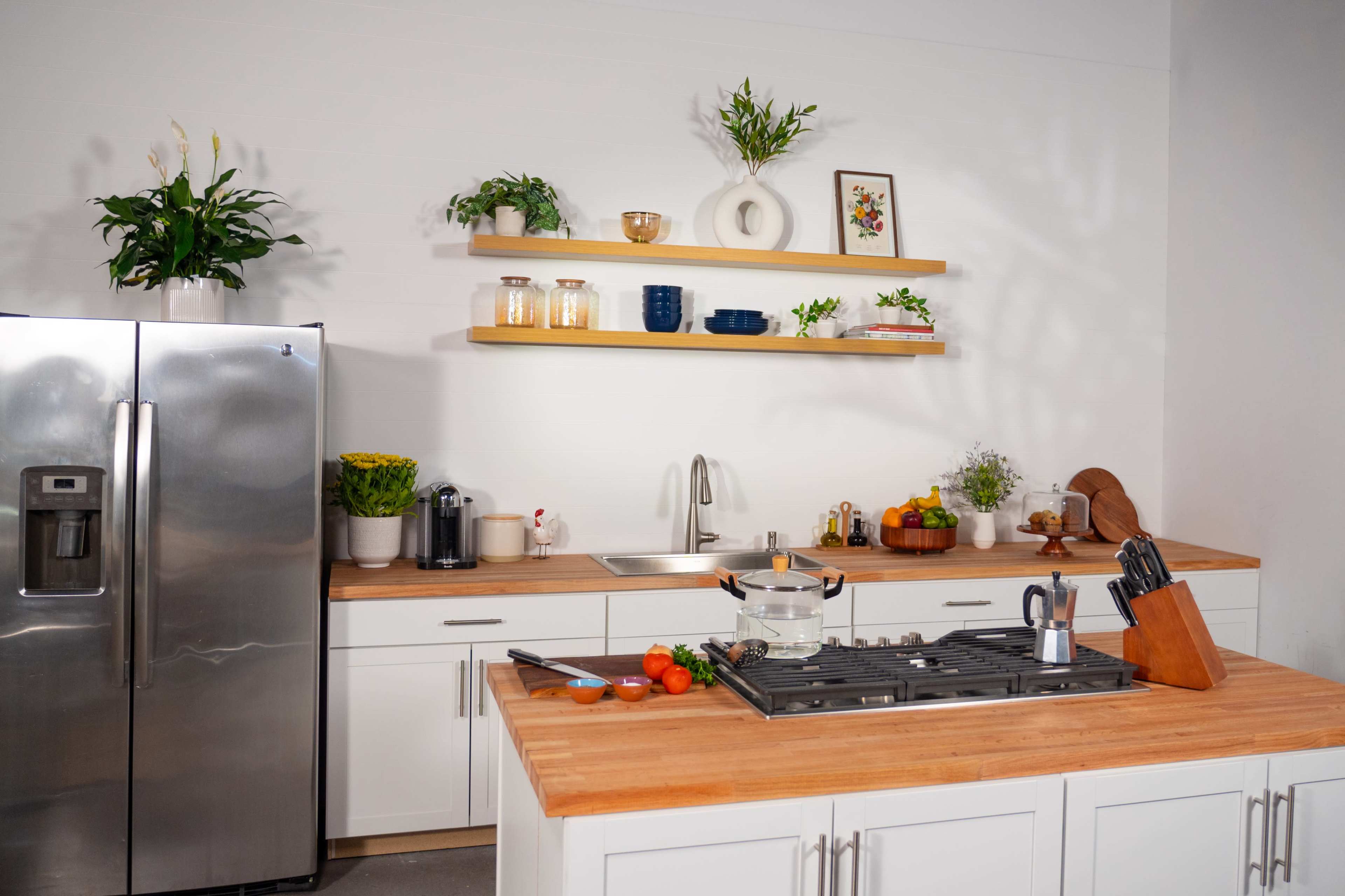 A modern kitchen features a sleek stainless steel refrigerator, wooden countertops, and open shelving with decorative items and potted plants.
