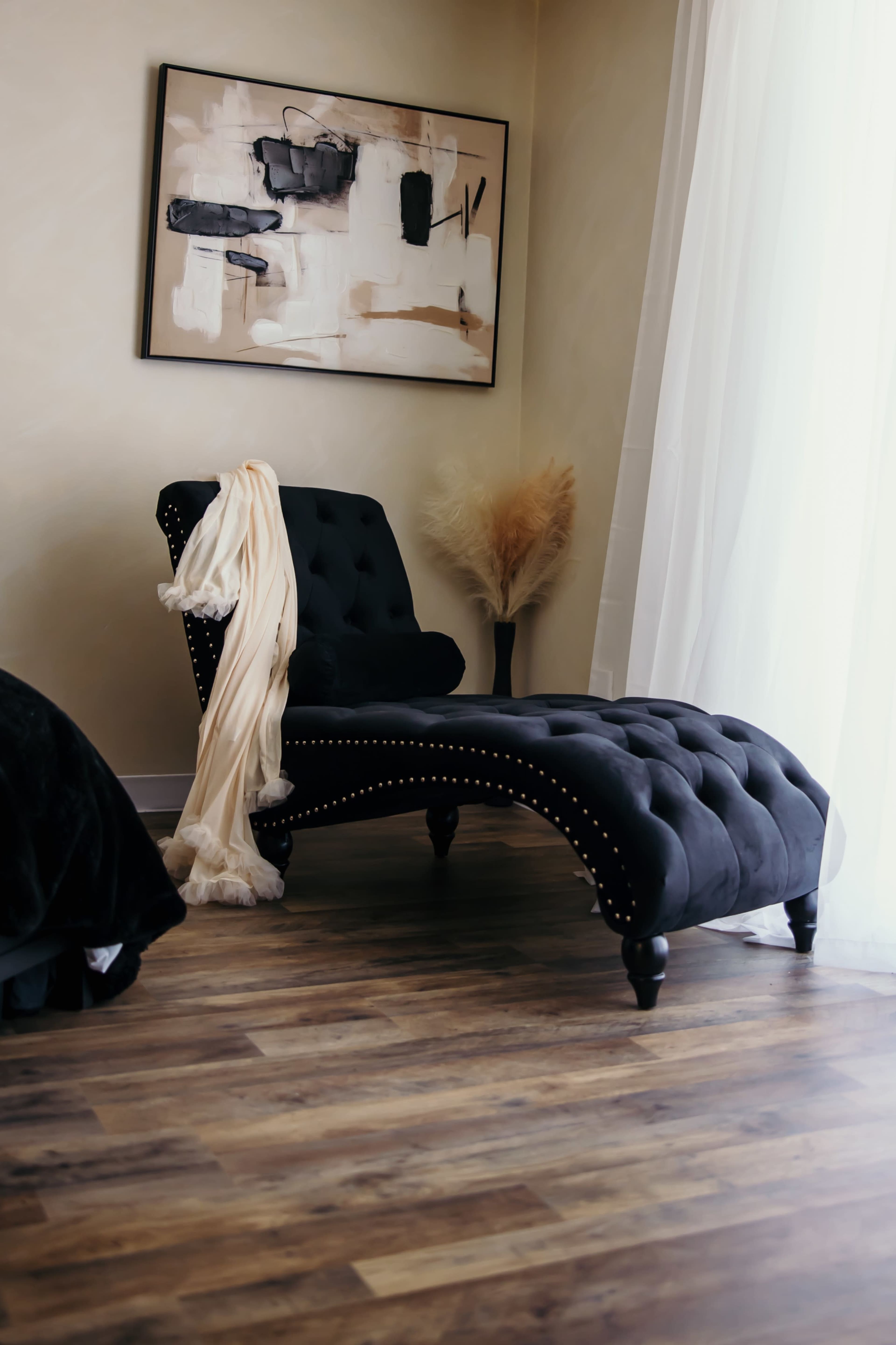 A black tufted chaise lounge with decorative nailhead trim is positioned beside a tall window, with a soft fabric draped over it and a decorative plant nearby.